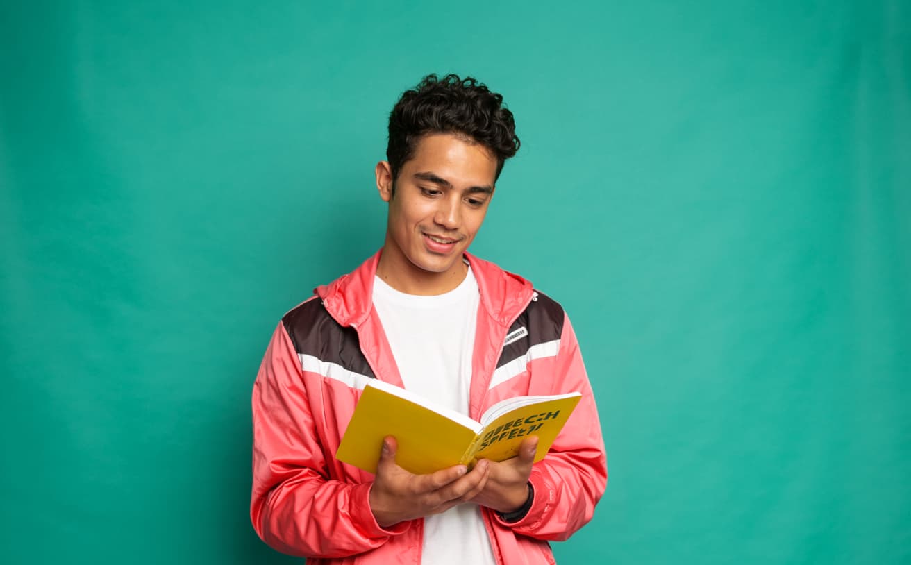 Young adult male standing against a teal background, reading a yellow book titled “Speech Speed” as part of his speech therapy journey.