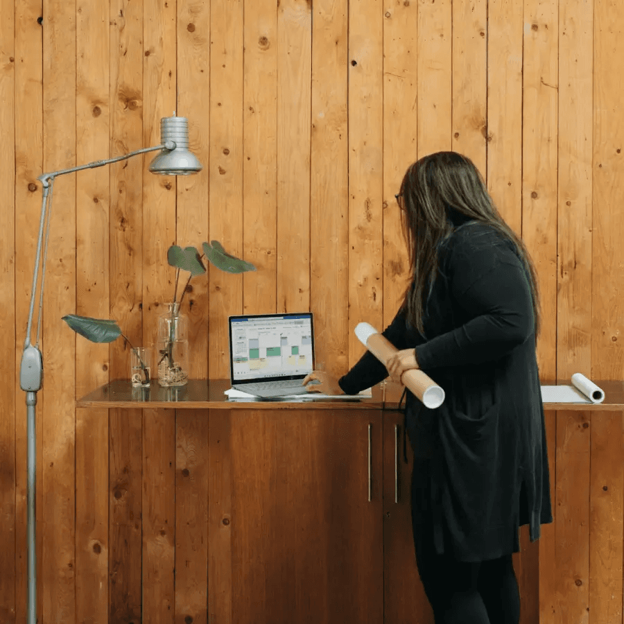 A person in a black sweater stands at a wooden console, arranging items with decorative plants nearby.