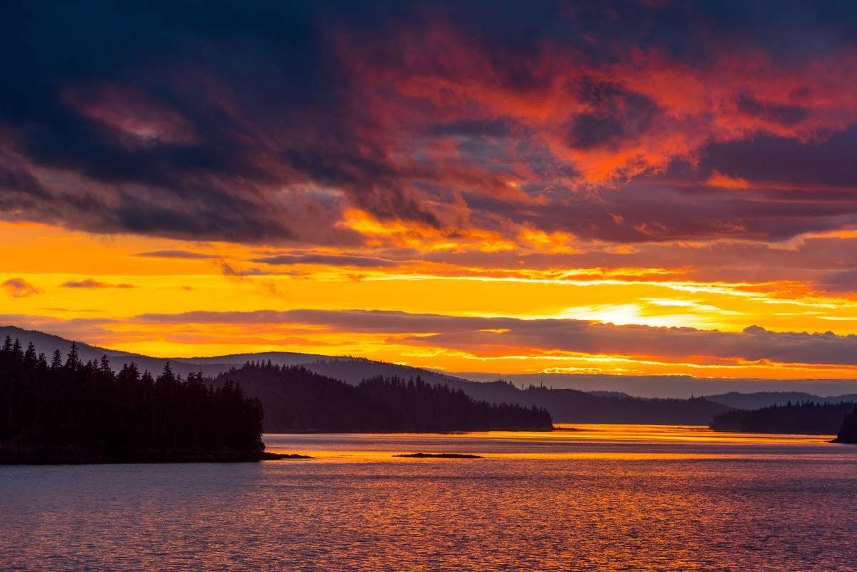 A sunset over a lake with dark pine trees in the foreground and orange and red clouds reflecting on the water.