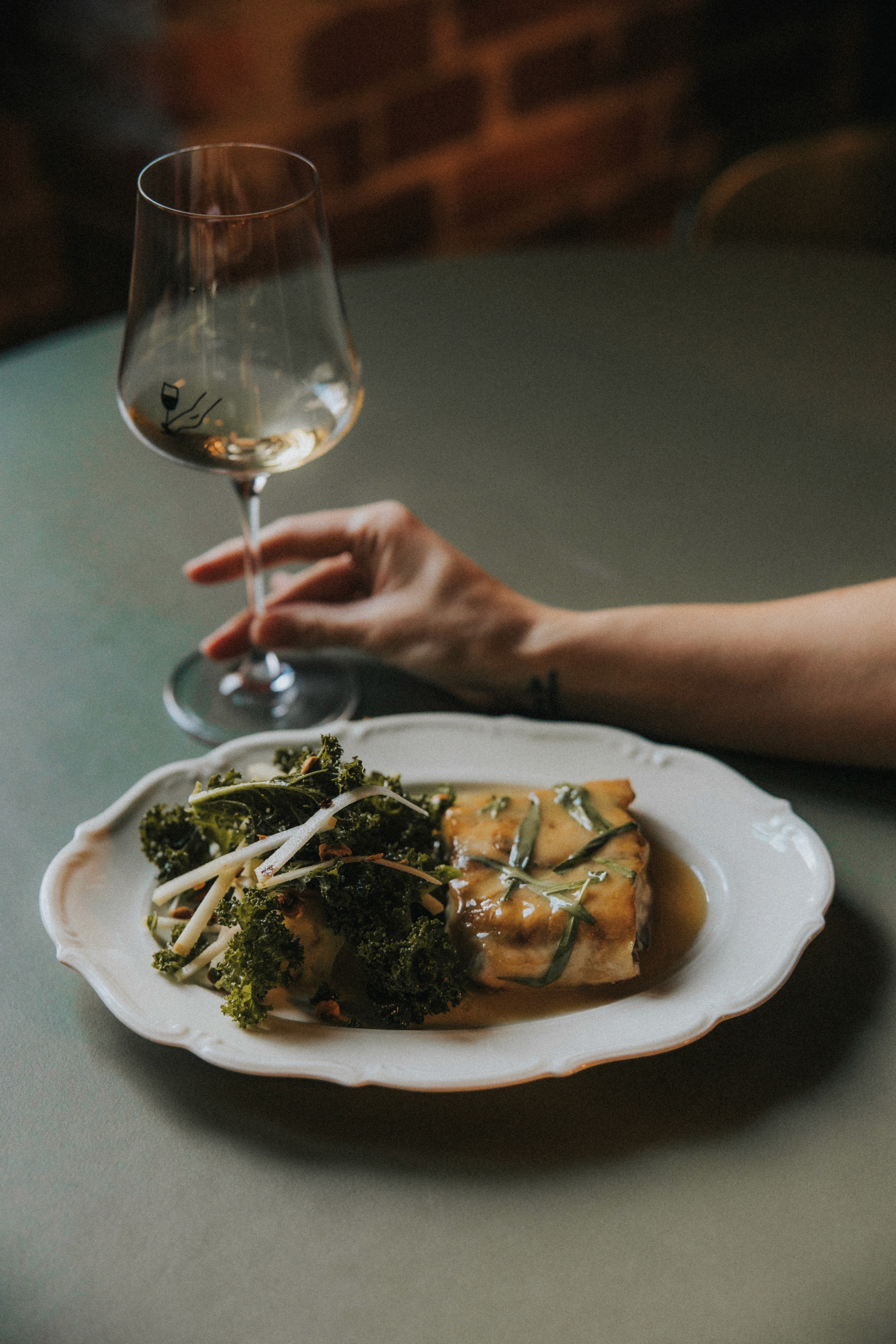 A hand holding a wine glass next to a plate of food, featuring greens and a golden-brown dish on a table.