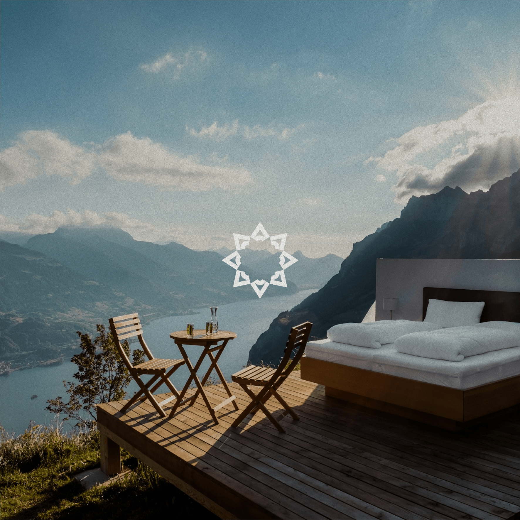 A wooden deck overlooks a scenic mountain landscape by a lake, featuring a cozy bed and a small table set with chairs, ideal for a serene outdoor experience.