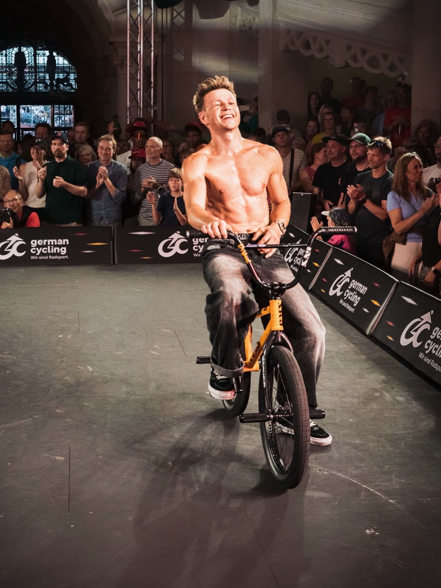 Chris Böhm sitzt lächelnd und oberkörperfrei auf seinem gelben BMX-Bike bei den BMX Finals, klatschendes Publikum und German Cycling-Banner im Hintergrund