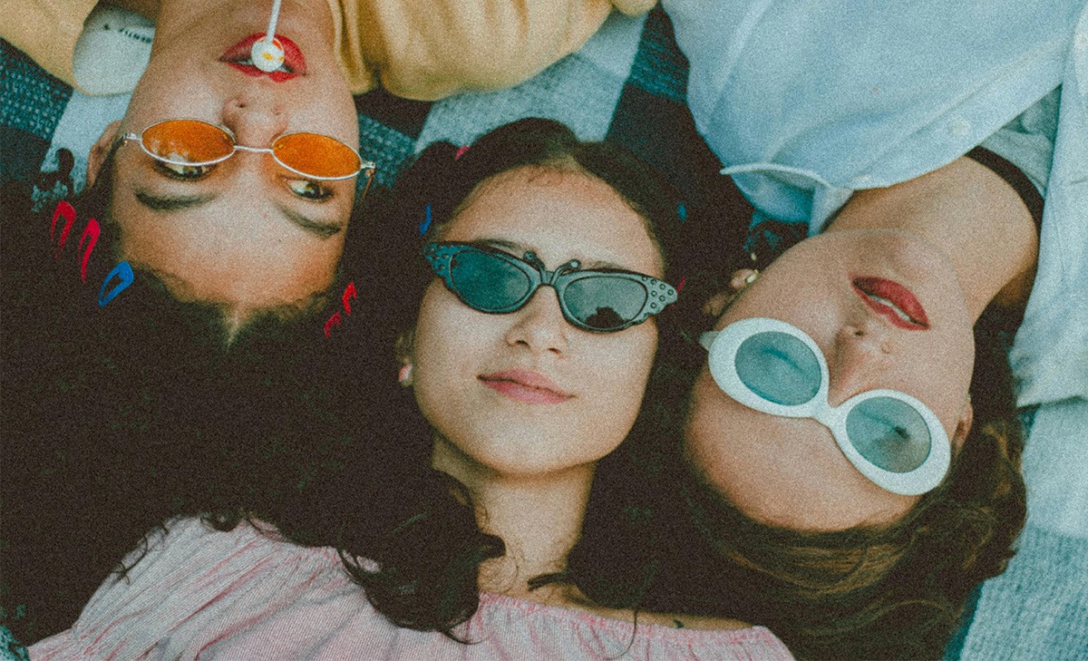 Three young women lying on a beach blanket wearing colorful vintage sunglasses, photographed from above in a nostalgic summer style.