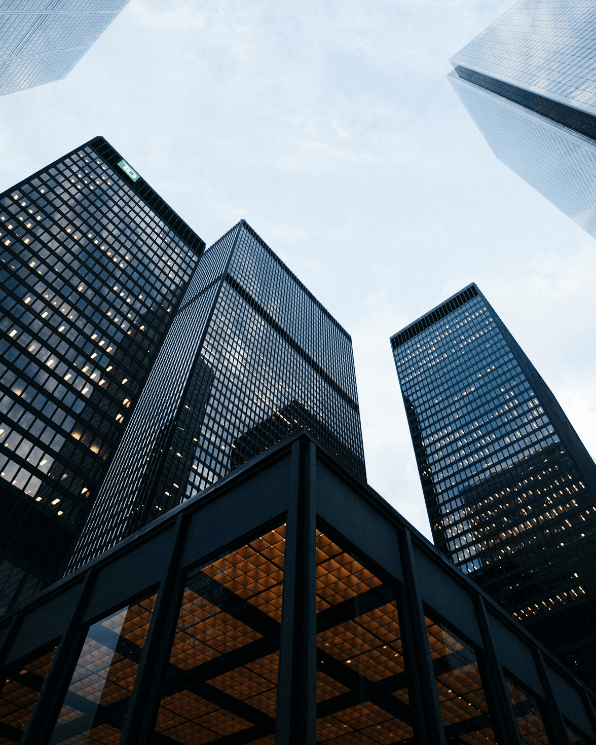 A stylish, modern lounge area featuring pastel green sofas, tan chairs, round wooden tables, and ceiling plants, with large windows and sheer curtains.