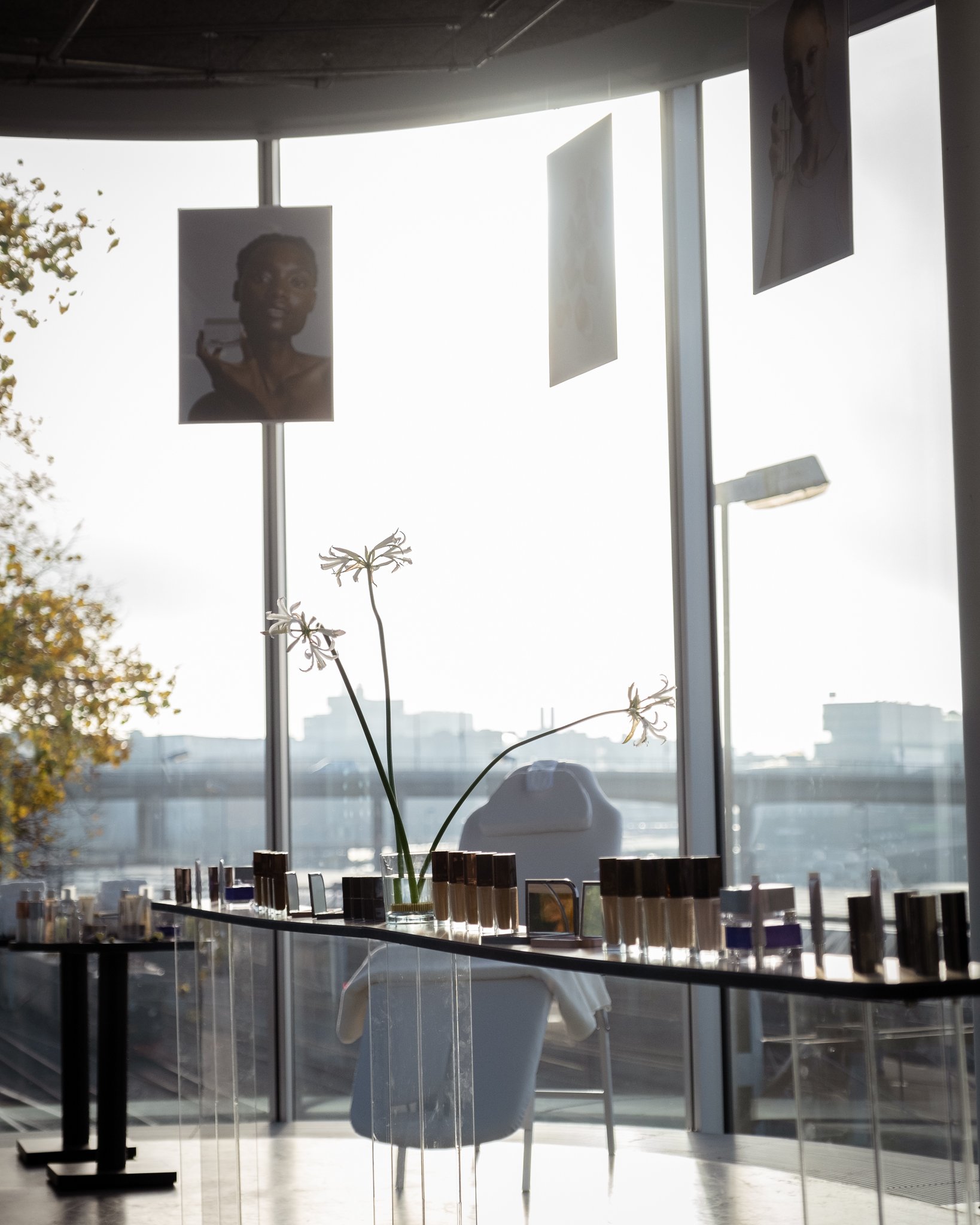 Interior scene with a large window showcasing a cityscape, a table with nail polish bottles, a single flower in a vase, and hanging photographs on the window.
