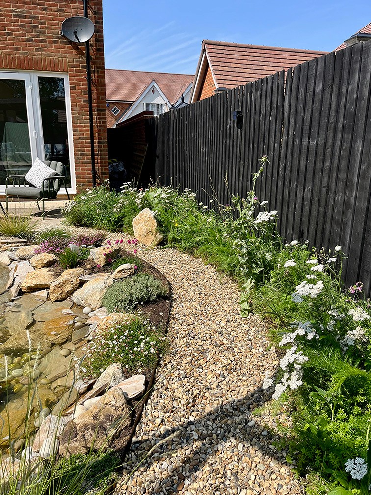 A sunny garden path lined with flowers leading to a house, bordered by a wooden fence.