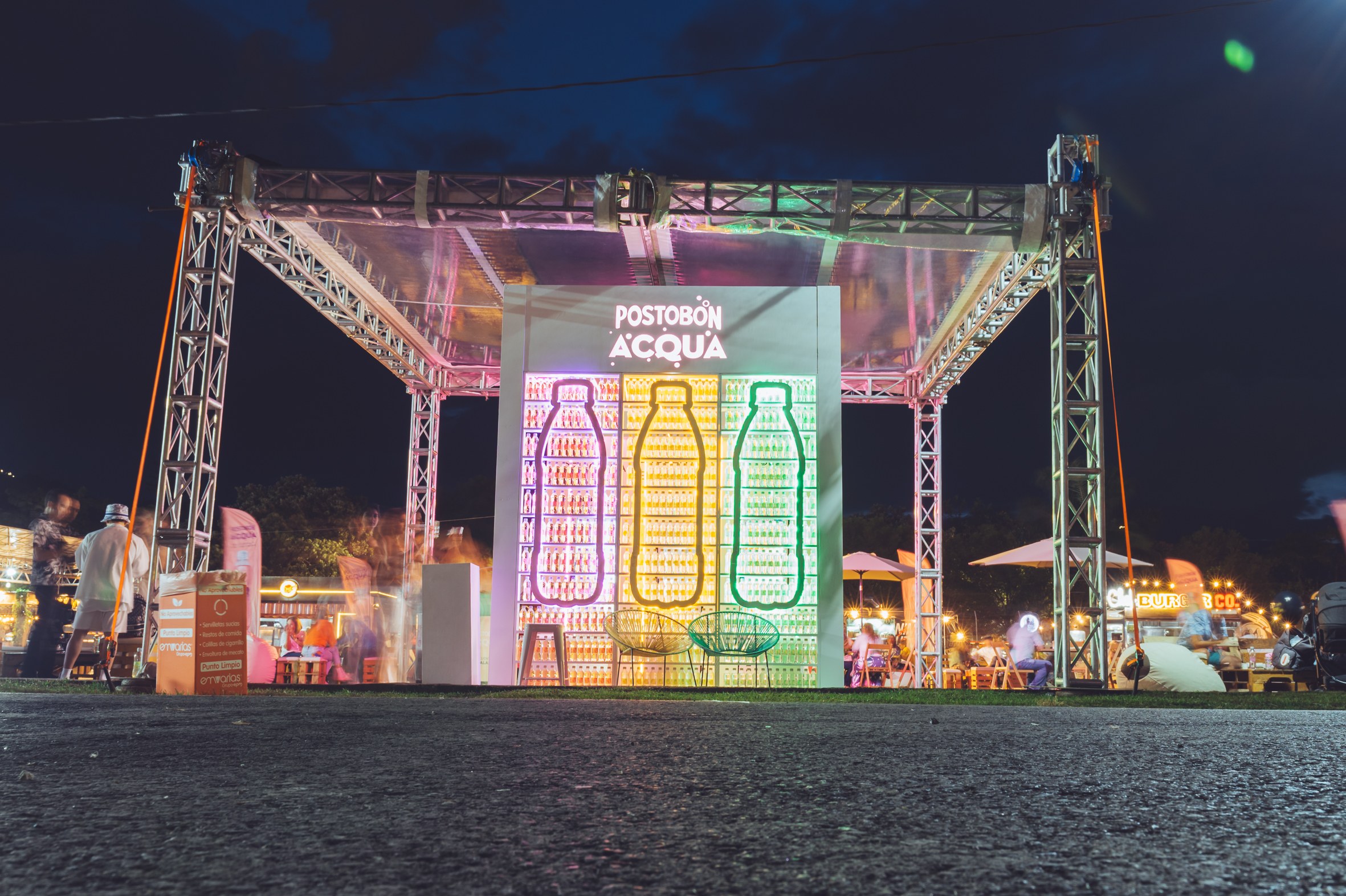 Estructura metálica iluminada del stand de Postobón ACQUA en Paradiso al anochecer, con botellas gigantes