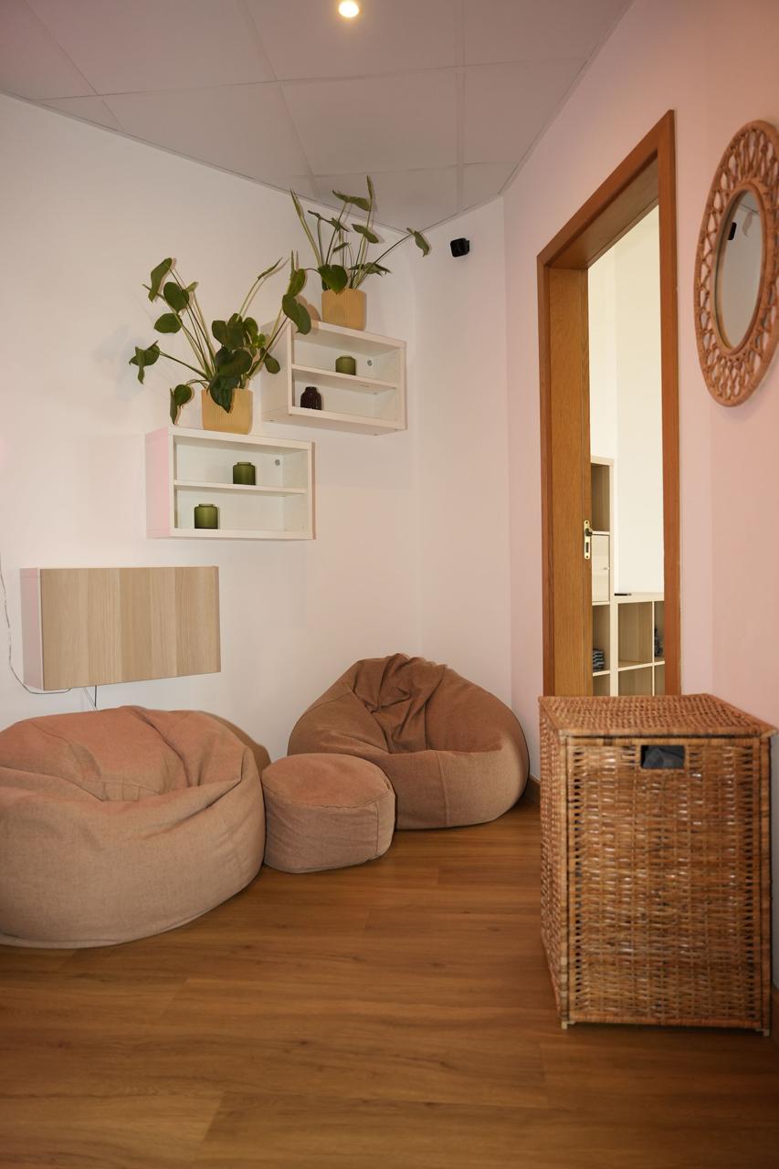 A cozy waiting area corner with comfortable beige bean bag chairs, a woven pouf, and white wall shelves decorated with green plants and candles.