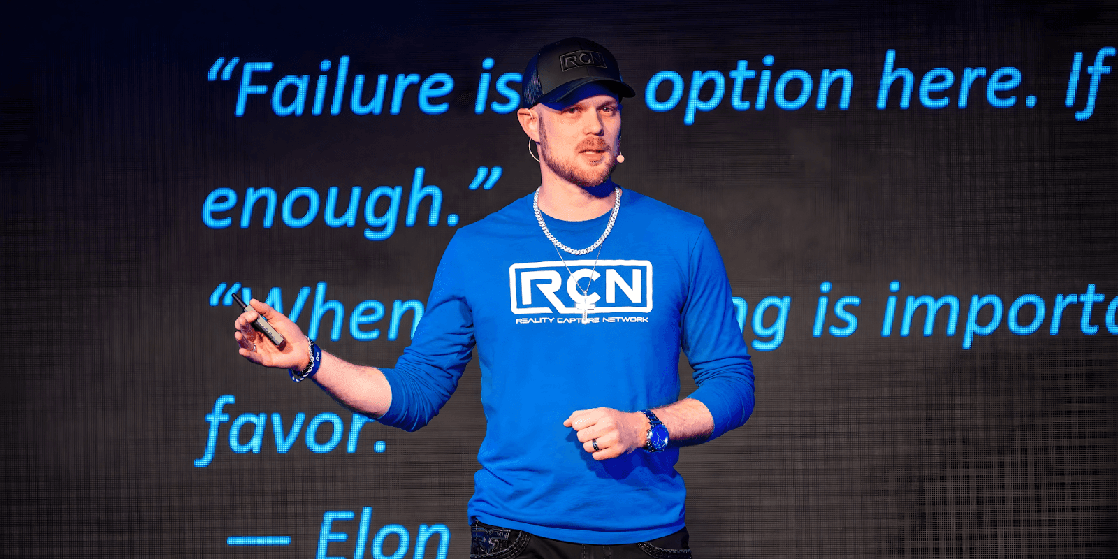 Matthew Byrd speaking at an event, wearing a blue Reality Capture Network shirt, with an inspirational quote on the screen behind.