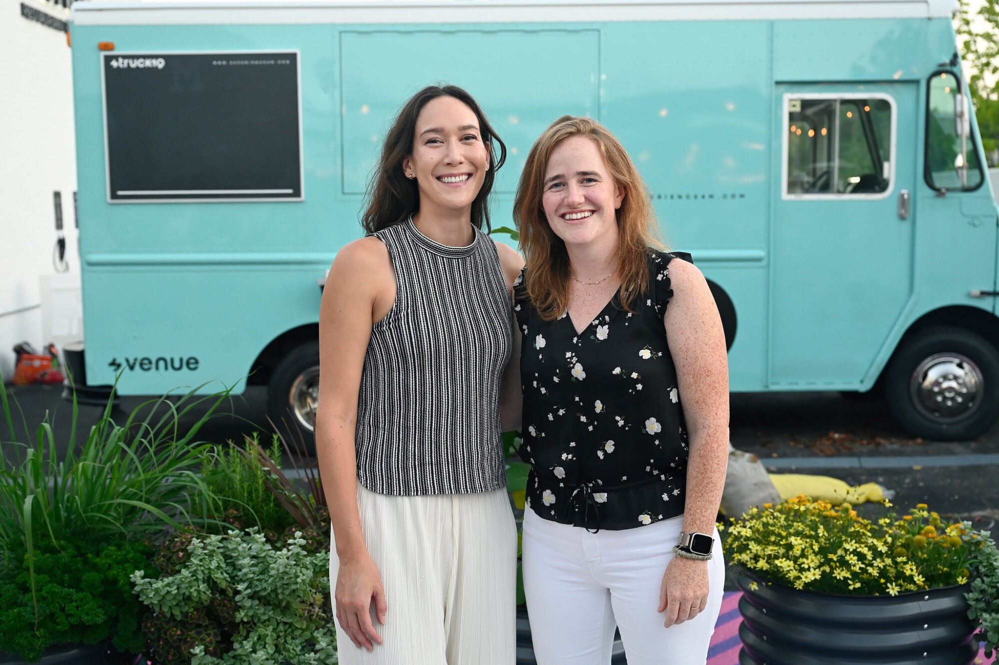 Two women smile in front of a teal food truck, surrounded by colorful flowers in a garden setting.