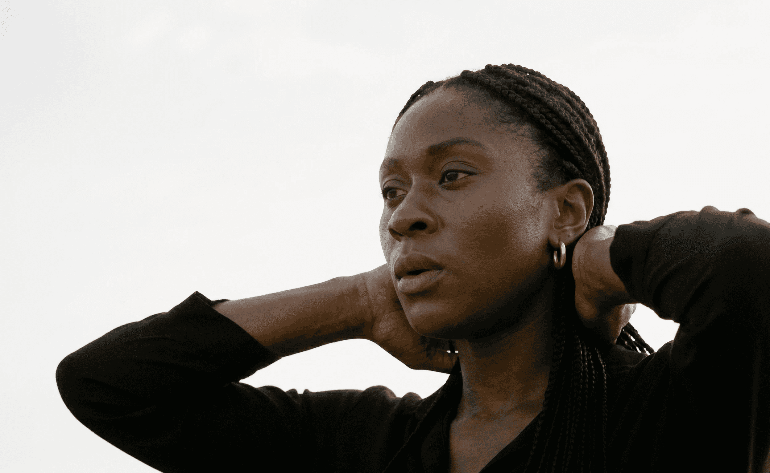 A person wearing a black top with braided hair is gazing thoughtfully off-camera against a neutral, pale background.