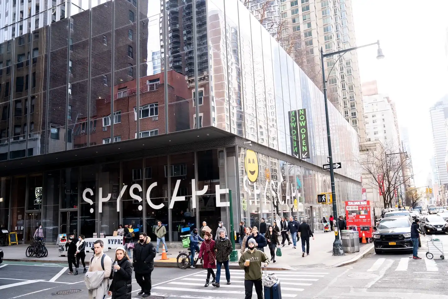 Busy New York street with crowds walking past the SPYSCAPE venue featuring large branding on the glass facade.
