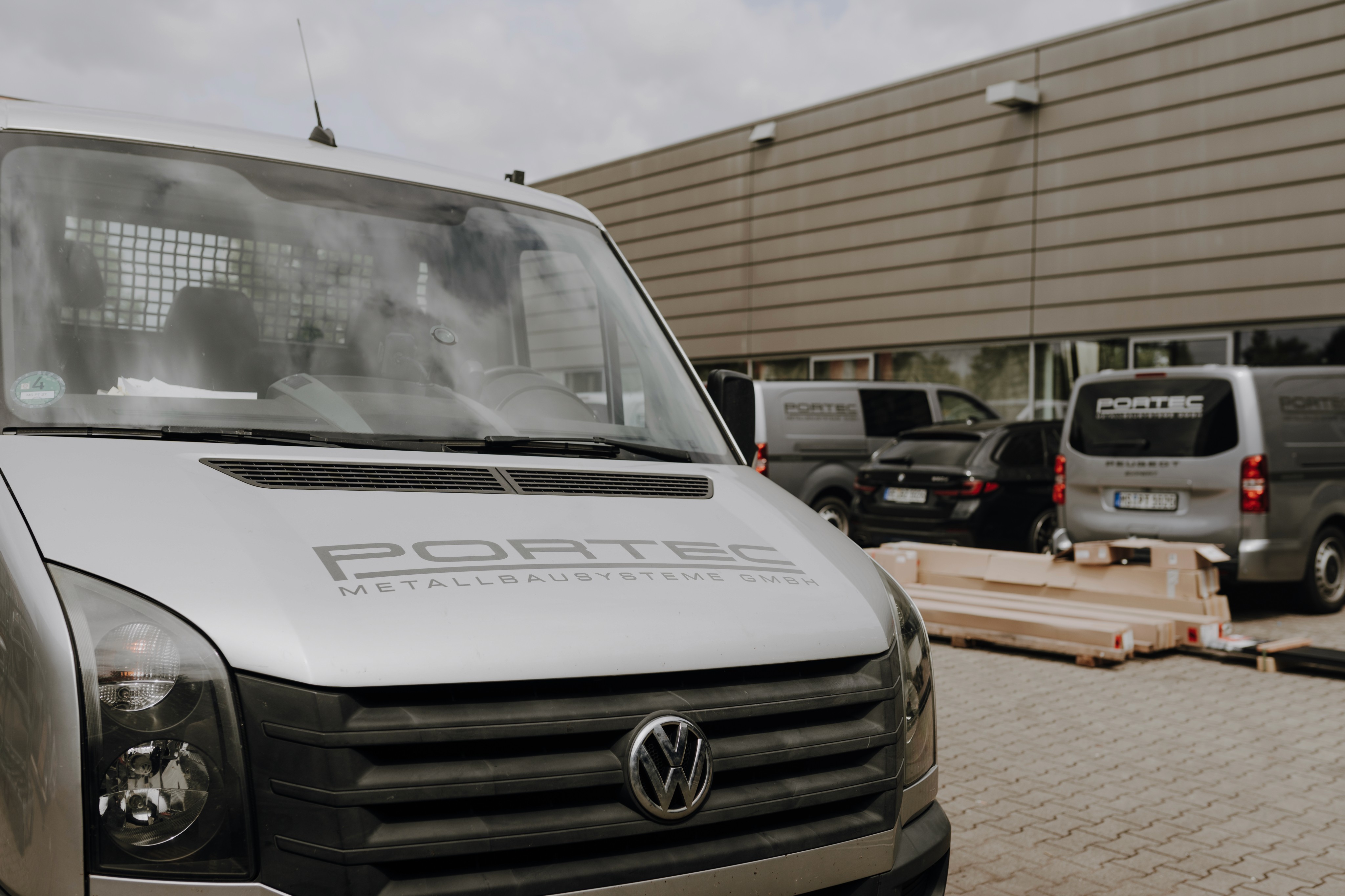 Close-up of a VW van with "PORTEC Metallbausysteme GmbH" logo parked outside a building. Other vans and a car in background.