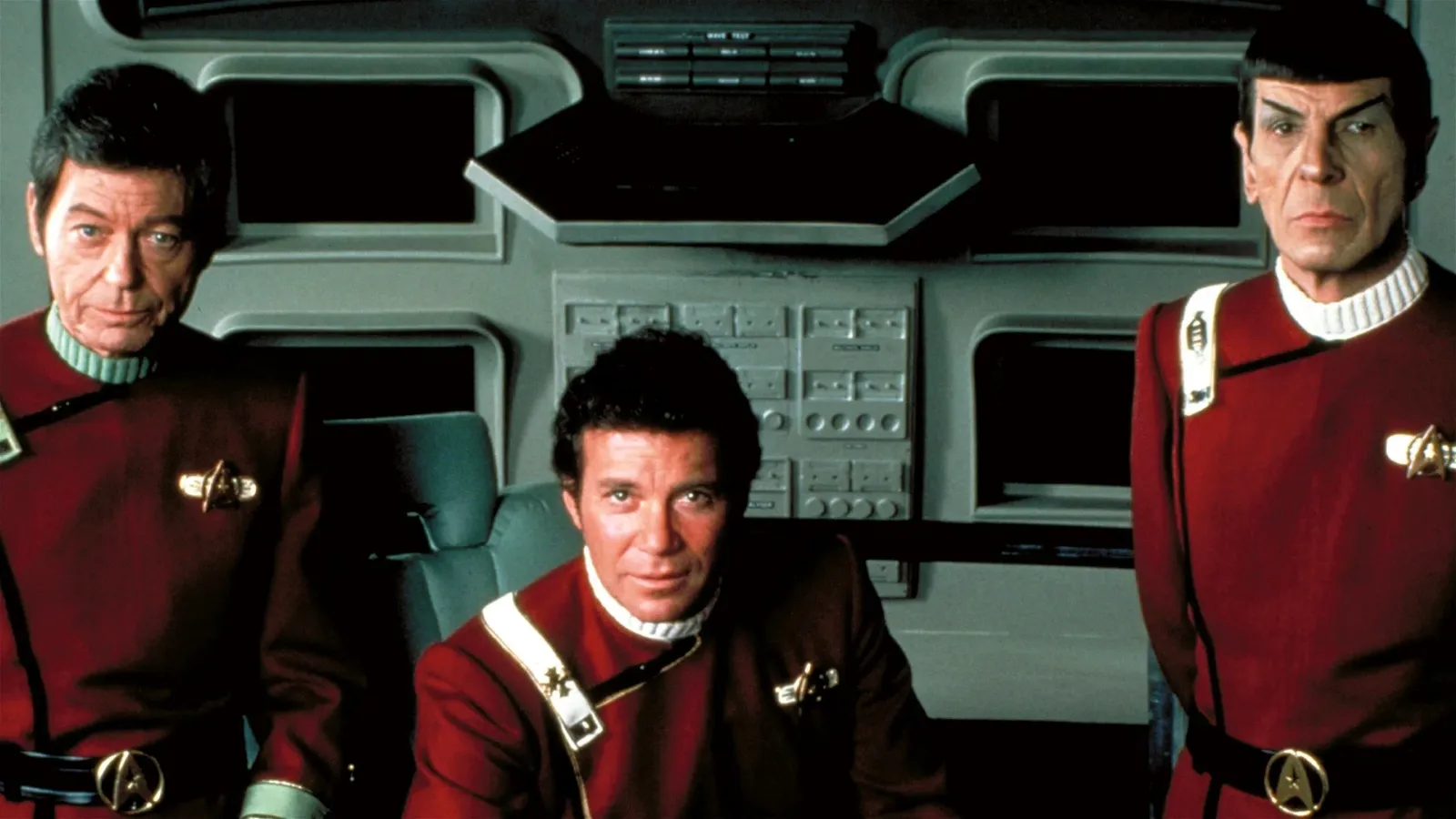 Three men in red and black sci-fi uniforms stand in what appears to be a futuristic spaceship control room, complete with control panels and screens in the background.