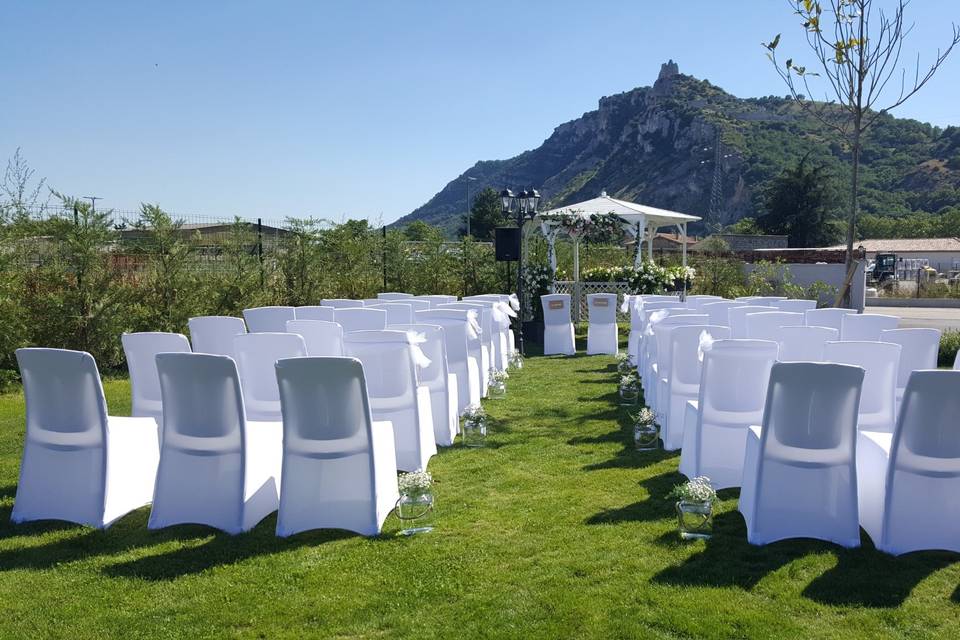 A serene outdoor wedding setup with white chairs arranged on green grass under a canopy of trees.