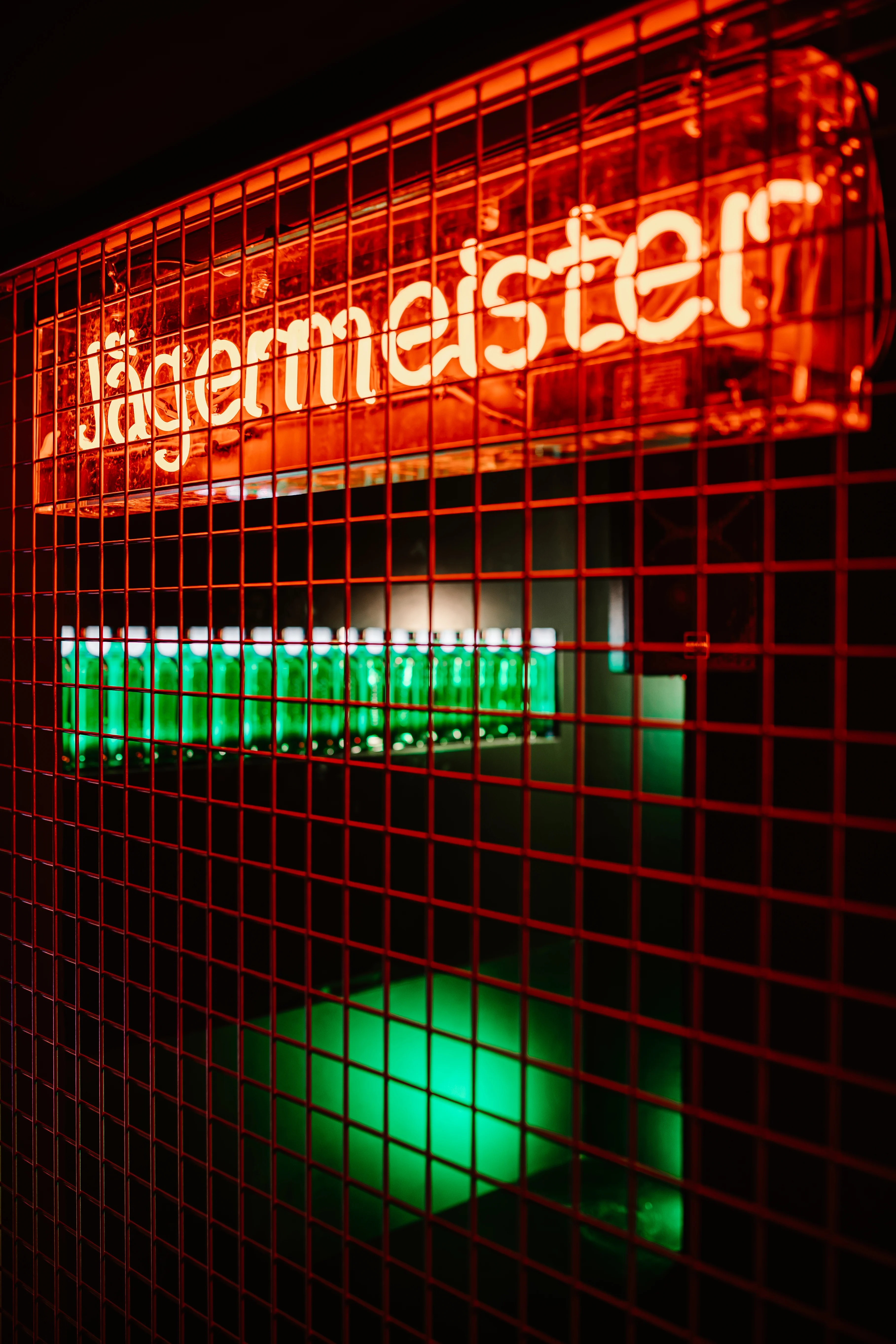 Orange Jägermeister neon sign behind a red metal grid at JÄGERMEISTER x CALL ME DRELLA, above a row of green backlit bottles in a wall niche.