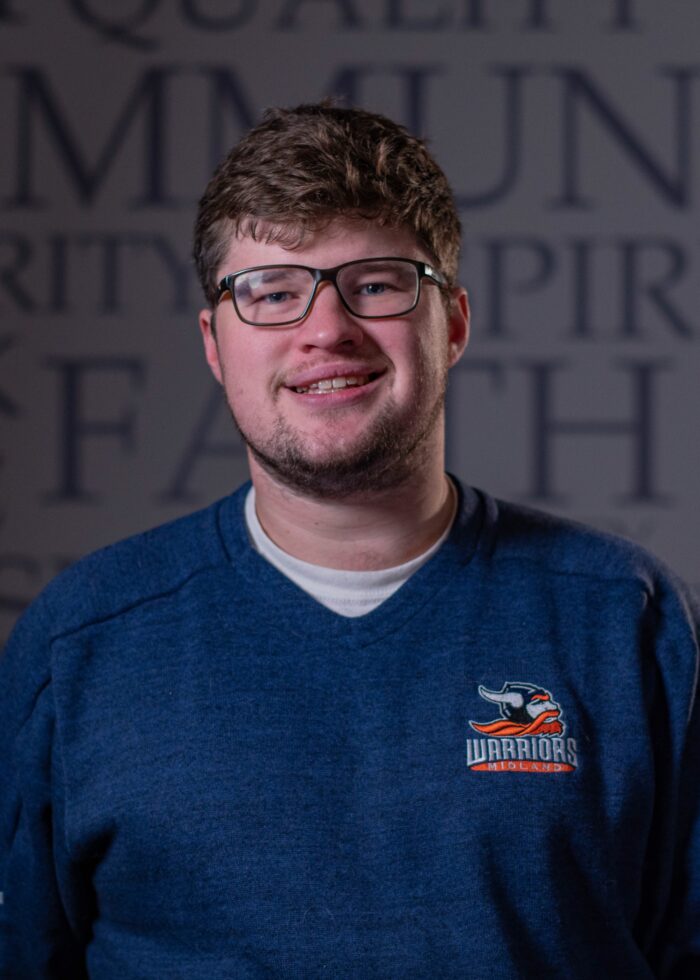 Smiling man wearing glasses and a blue Warriors sweatshirt poses in front of a blurred background with large text reading “COMMUNITY,” “SPIRIT,” and “FAITH.”