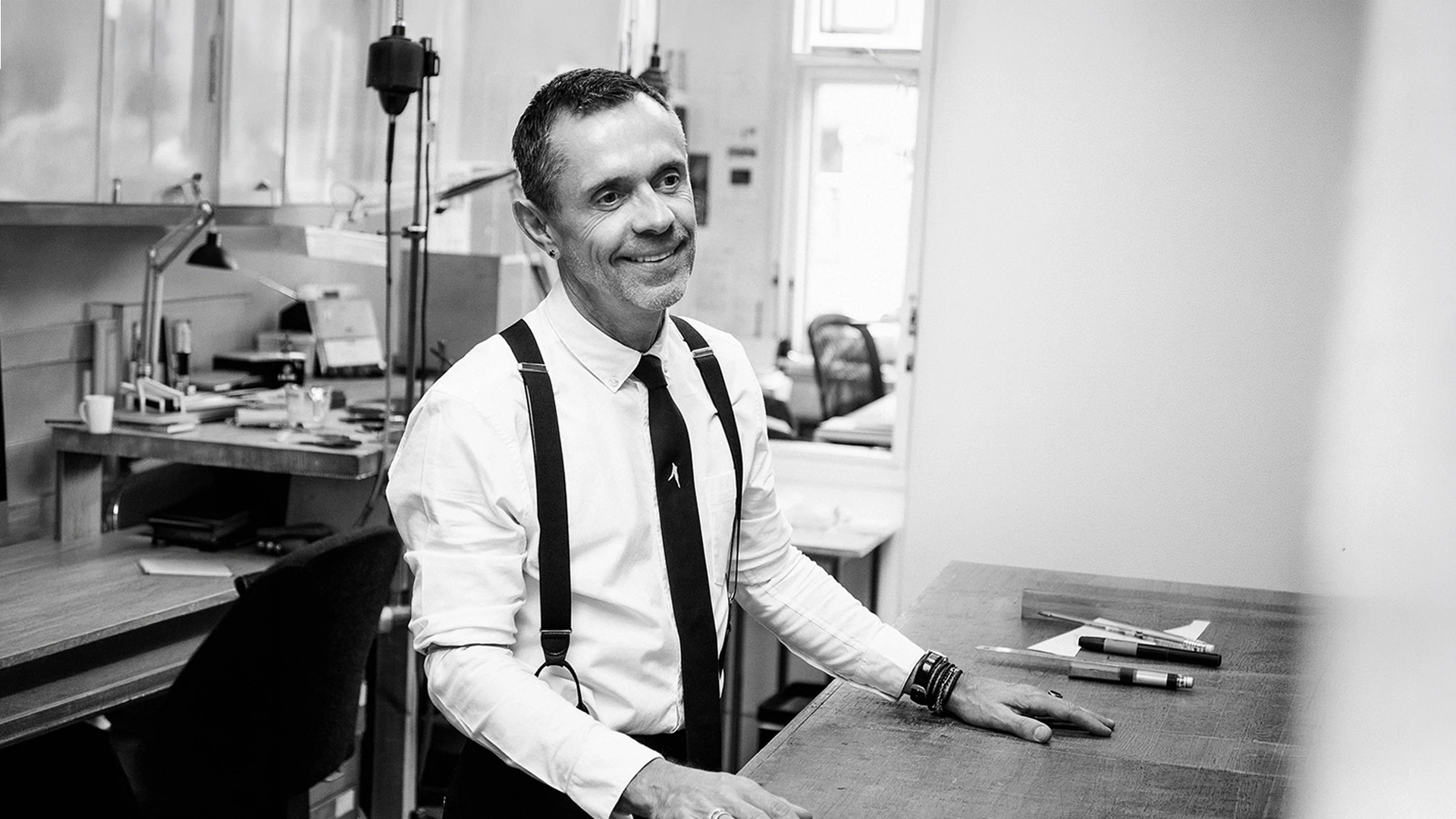 Black and white portrait of jewelry designer Shaun Leane in his workshop studio, wearing braces and leaning on a workbench.
