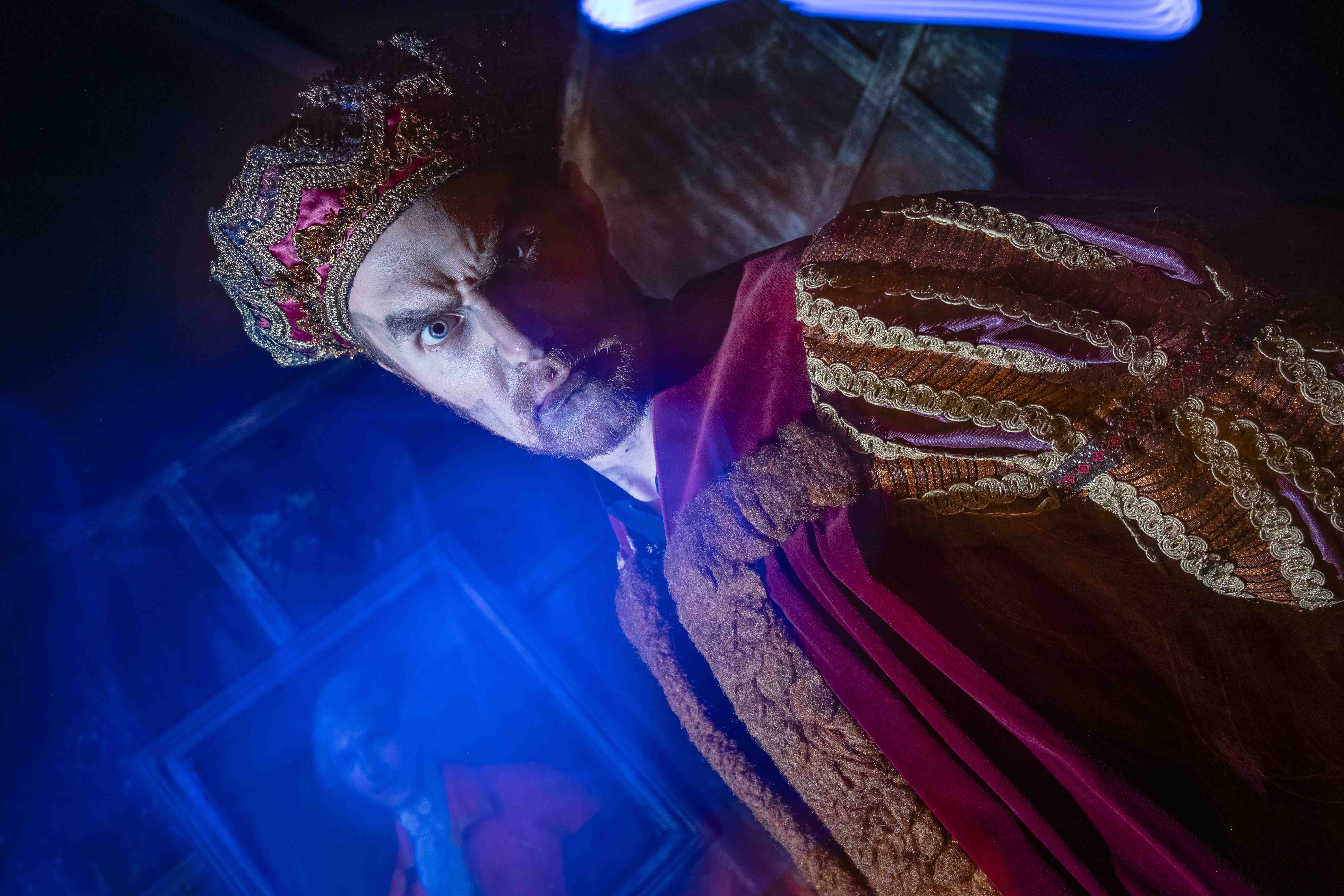 A man dressed in rich, ornate vintage royal attire stands under dramatic blue lighting, wearing a jeweled crown and embroidered robe, exuding a regal and intense expression.