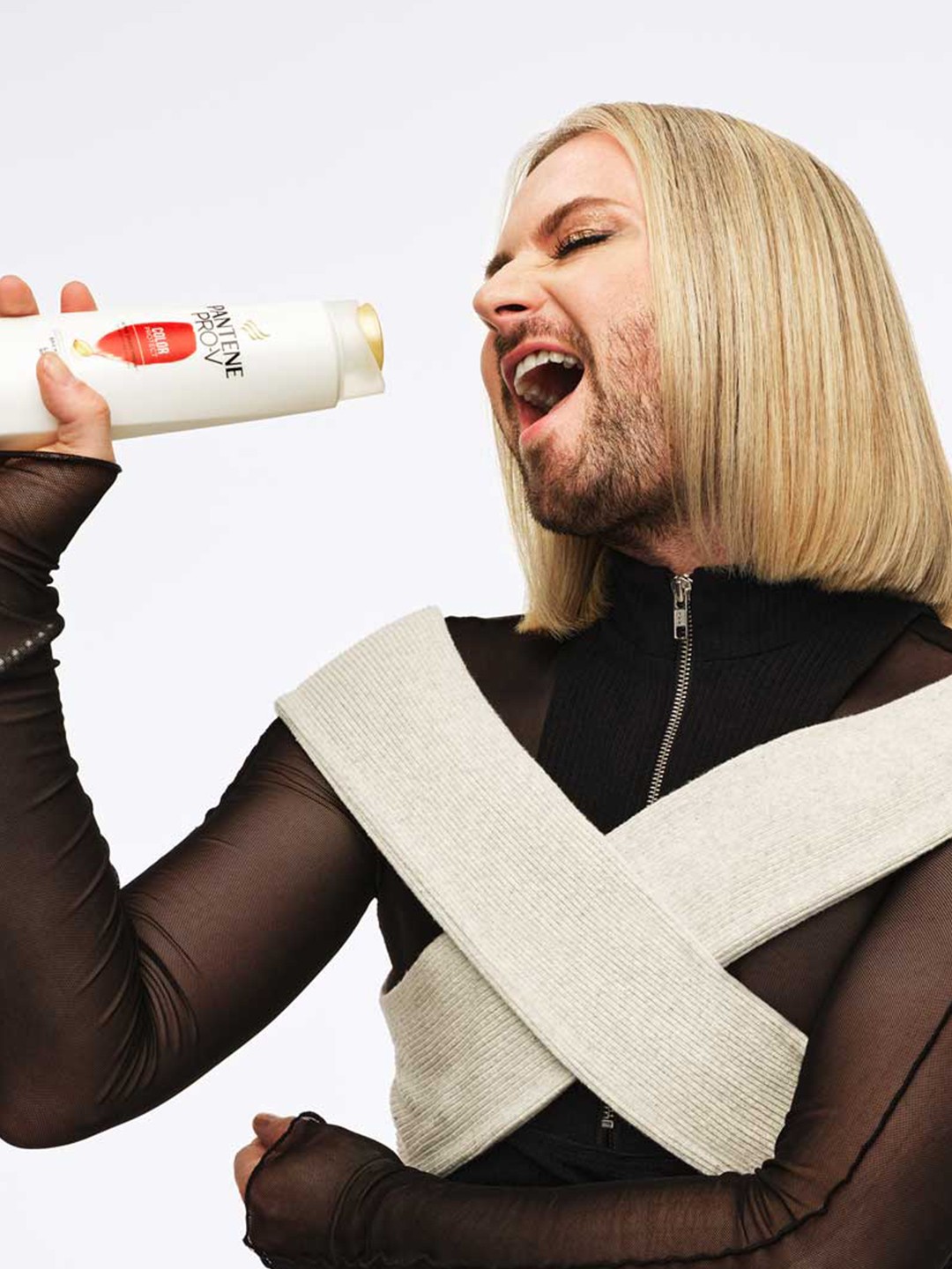 A person with shoulder-length blonde hair poses with one arm raised, wearing a sheer black top.