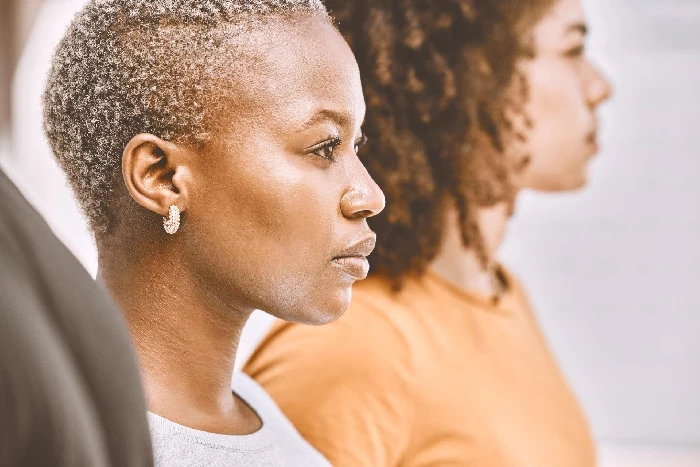 A imagem mostra duas mulheres em perfil, ambas com expressões sérias e concentradas, olhando para a frente. A mulher à esquerda tem o cabelo curto e usa um brinco, enquanto a mulher à direita tem o cabelo cacheado e veste uma camiseta de cor laranja. A composição da imagem foca nos rostos das mulheres, transmitindo uma sensação de determinação e foco. A iluminação suave destaca os contornos de seus rostos e adiciona uma atmosfera contemplativa e introspectiva à cena.