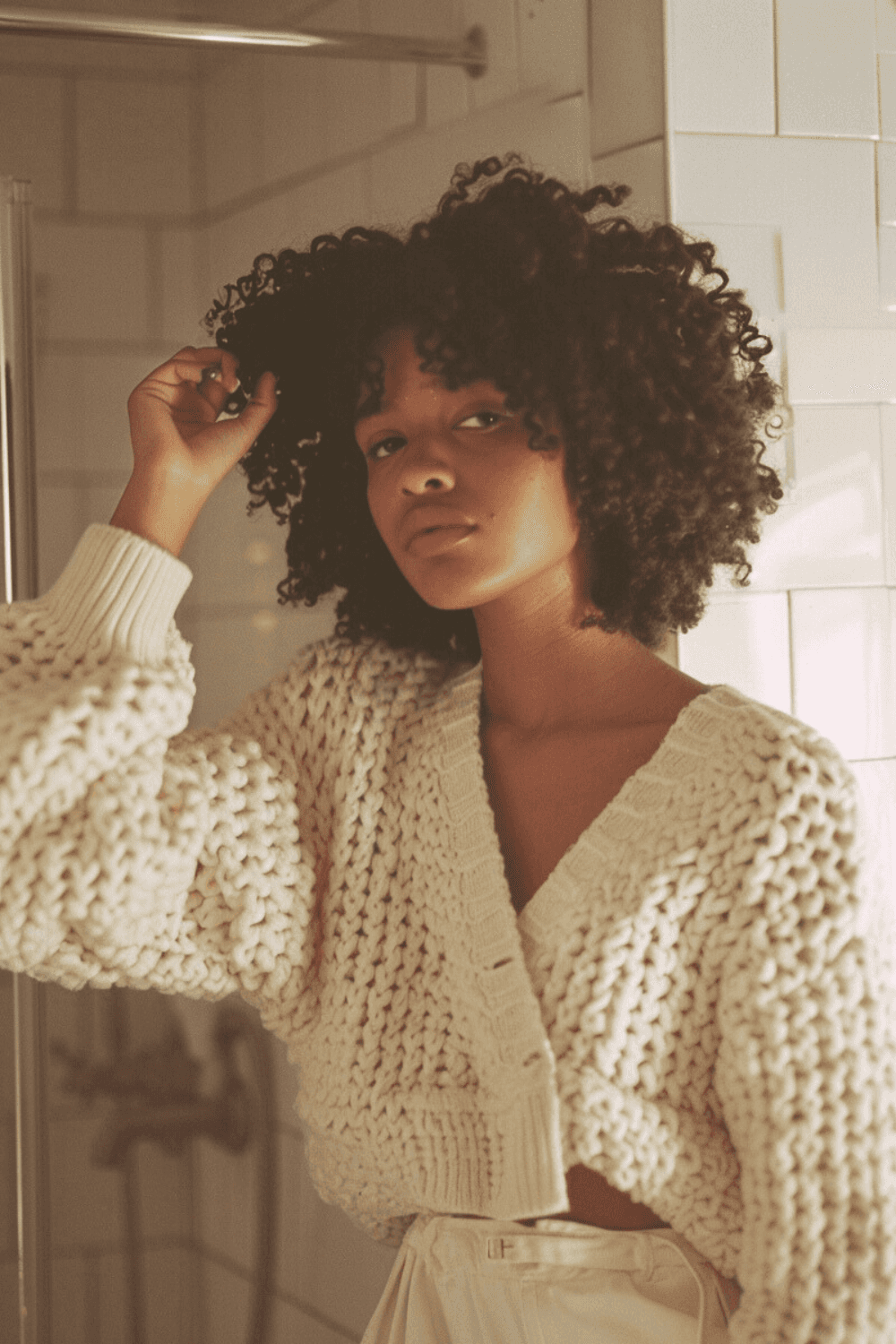 Postpartum mother looking at her reflection while touching her natural hair, noticing hair changes after birth in soft morning bathroom light