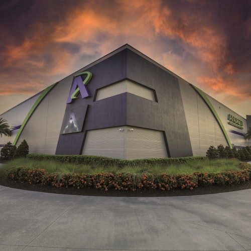 Голяма, модерна сграда със зелени акценти под драматично, оранжево и лилаво небе, заобиколена от озеленени храсти.