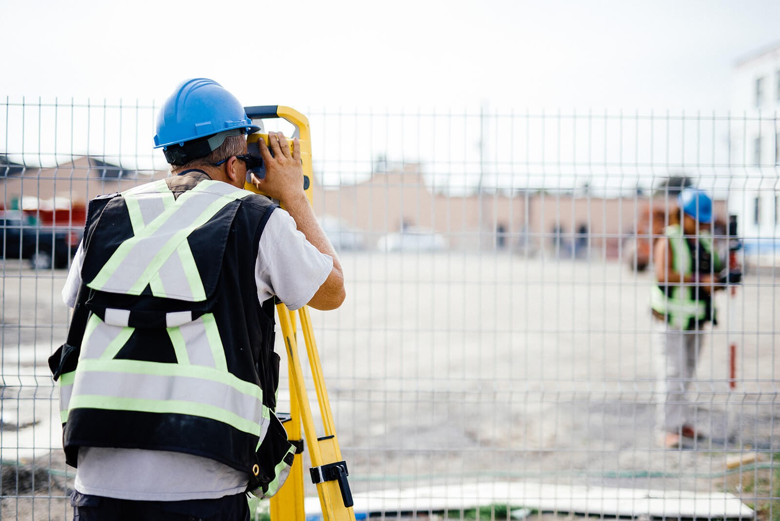 A construction worker in a yellow hard hat and safety goggles uses a drill on wood outside a wooden house under a clear blue sky.