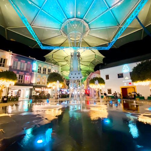 A brightly lit outdoor plaza at night with a large canopy, illuminated buildings, trees with string lights, and fountains on wet pavement.
