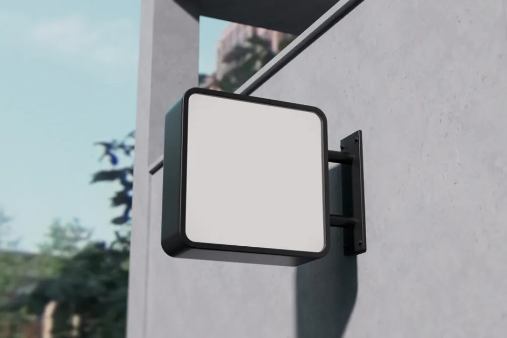 Square wall-mounted signage mockup with a black frame, attached to a concrete wall. The blank sign faces outward in natural lighting, with a blurred urban background.
