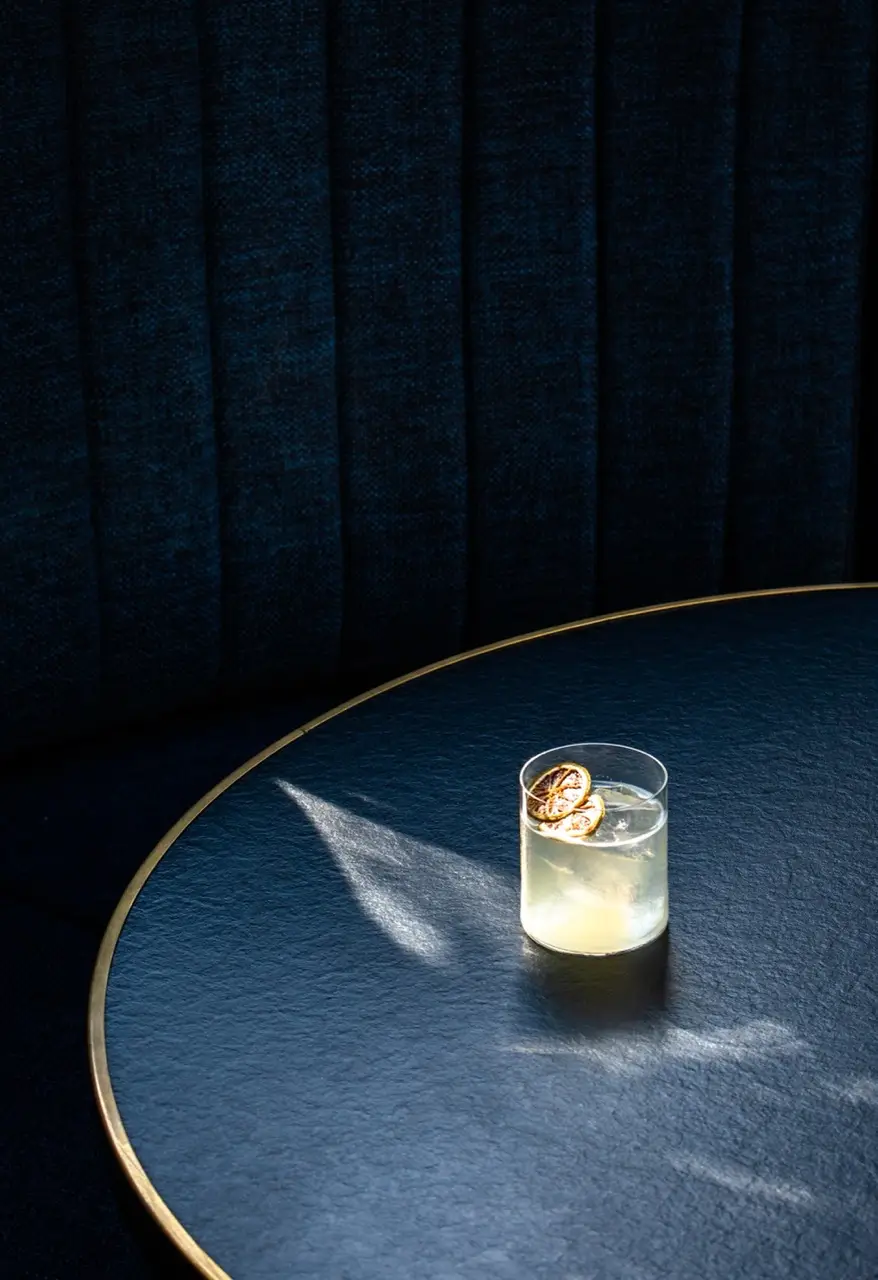 A solitary cocktail glass with a light-colored drink garnished with a thin lemon slice sits on a dark, round table bathed in soft sunlight.