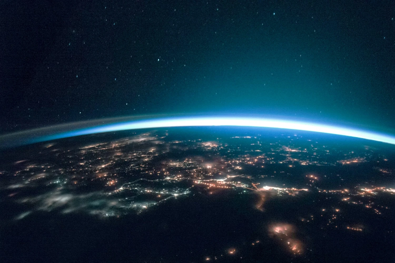 View of Earth from space at night, showcasing glowing city lights, the curved horizon, and a faint blue atmosphere under a starry sky.