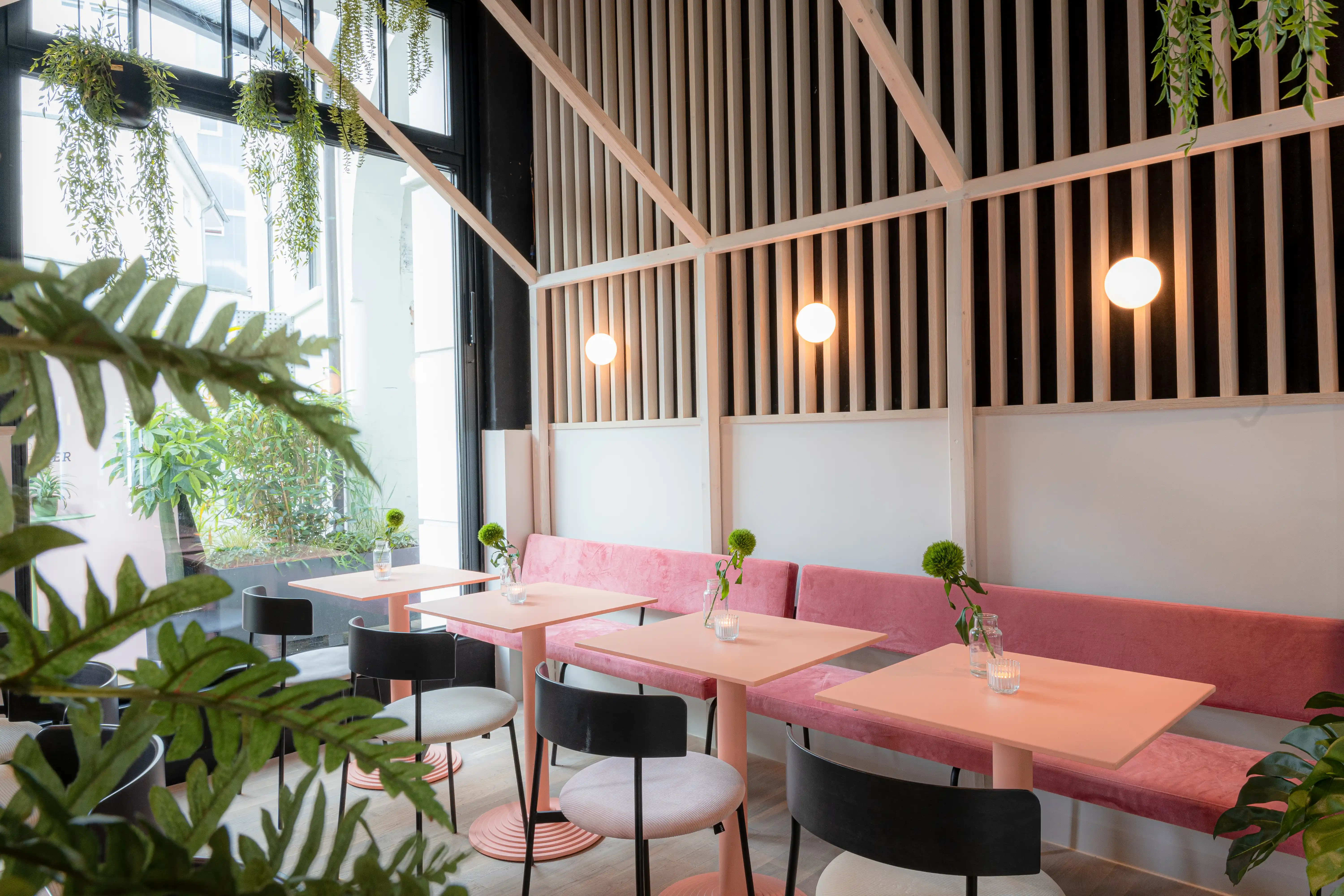 Window seating at LOKAL VIER with a pink velvet banquette, pink tables, black-and-cream chairs, white globe lights and a wooden slat framework above.