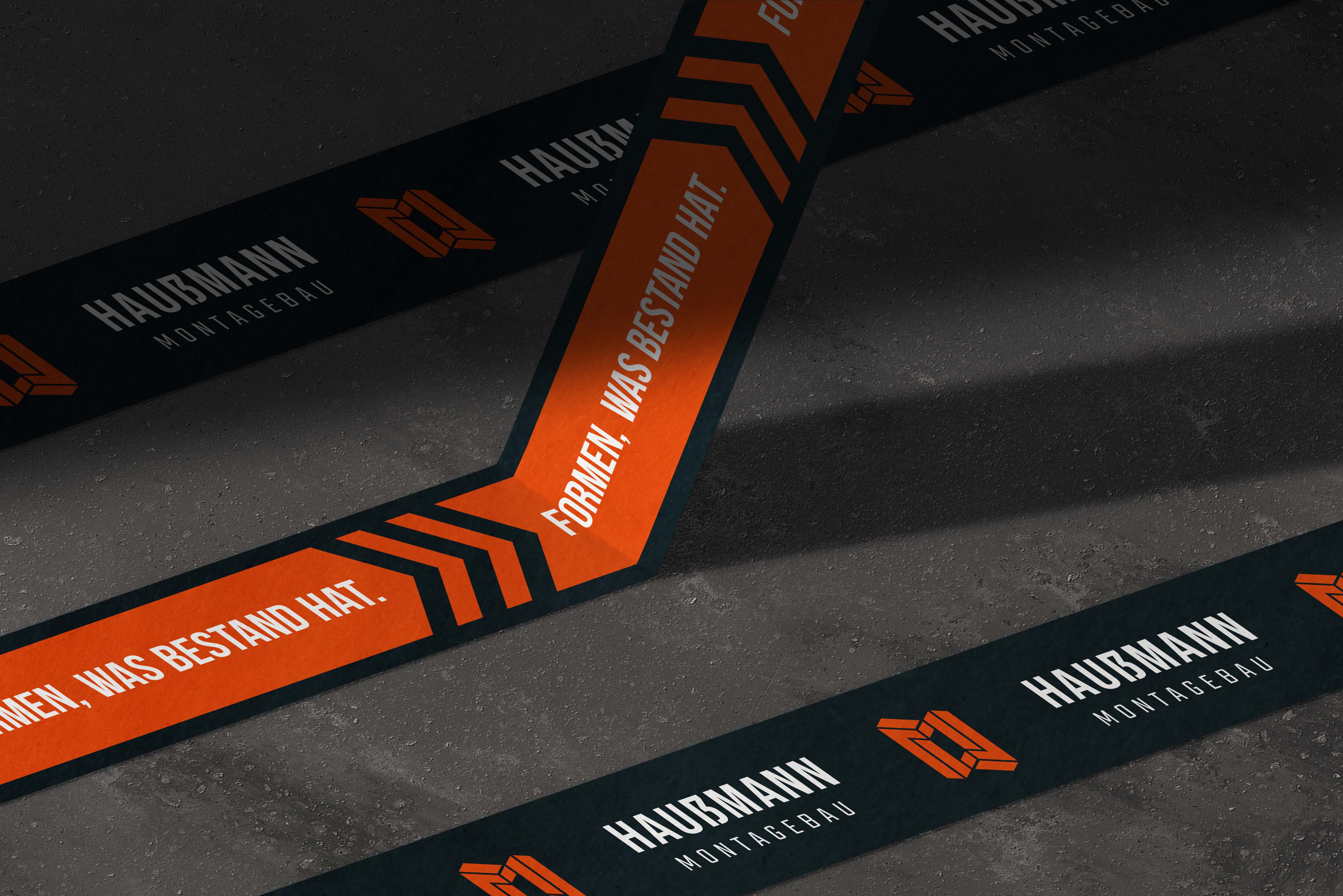 Schwarzes und orangefarbenes Klebeband mit der Aufschrift „Formen, was Bestand hat“ und dem Logo von Haussmann Montagebau auf grauem Boden.