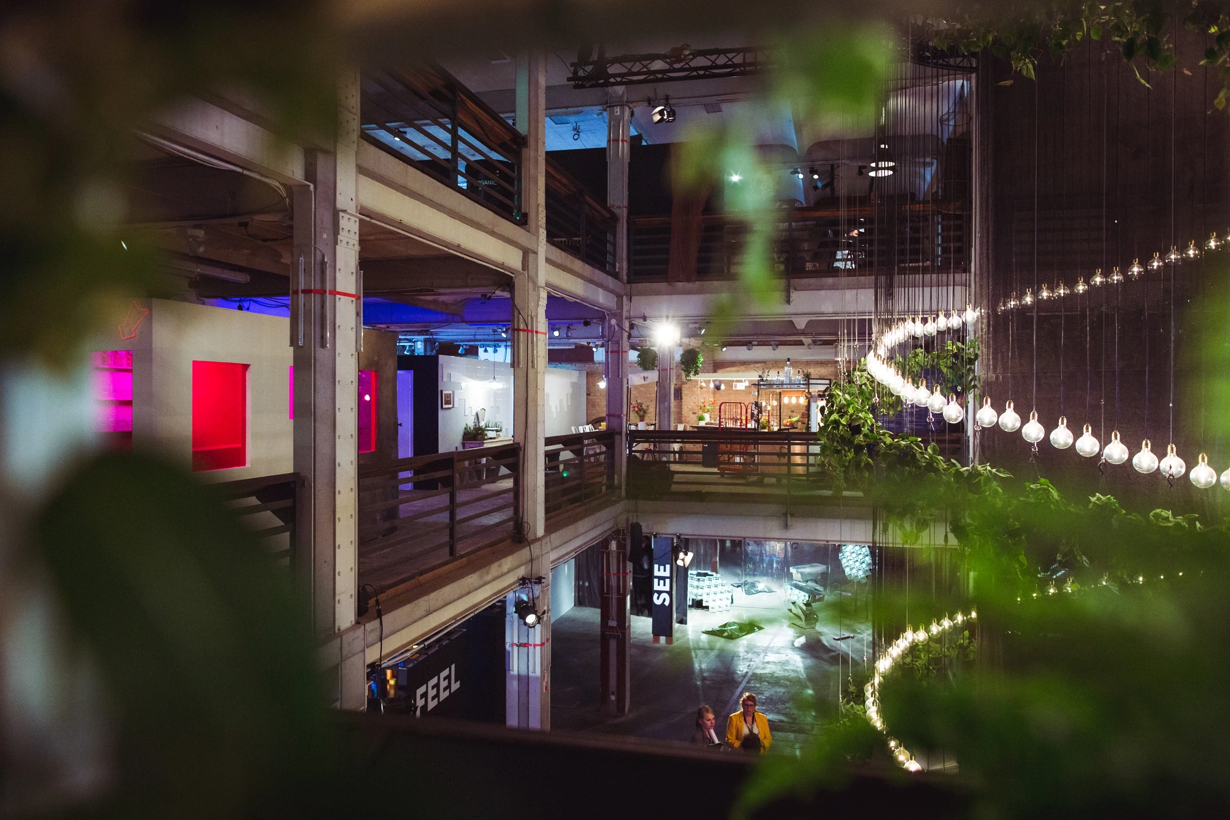 View across the multi-level DIAGEO BCB18 event space with FEEL and SEE signs, light garlands, greenery and exhibition areas on concrete galleries.