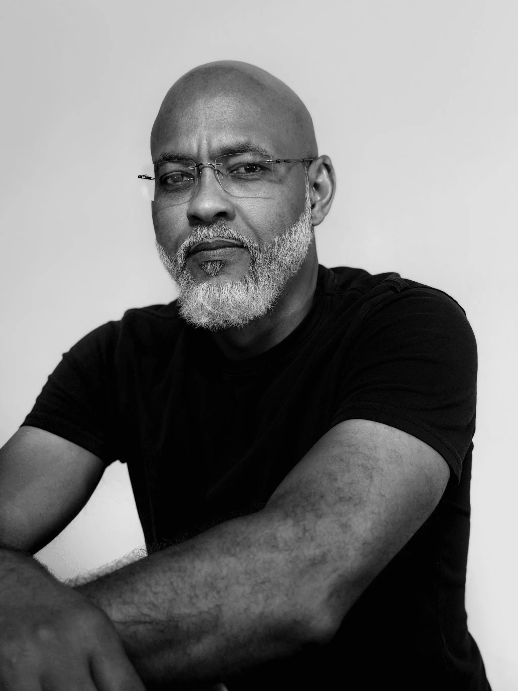 Photo of Kevin James, he has a gray beard and shaved head. He is wearing glasses and a black t-shirt and is posing with his arms crossed against a neutral background in this black and white portrait.