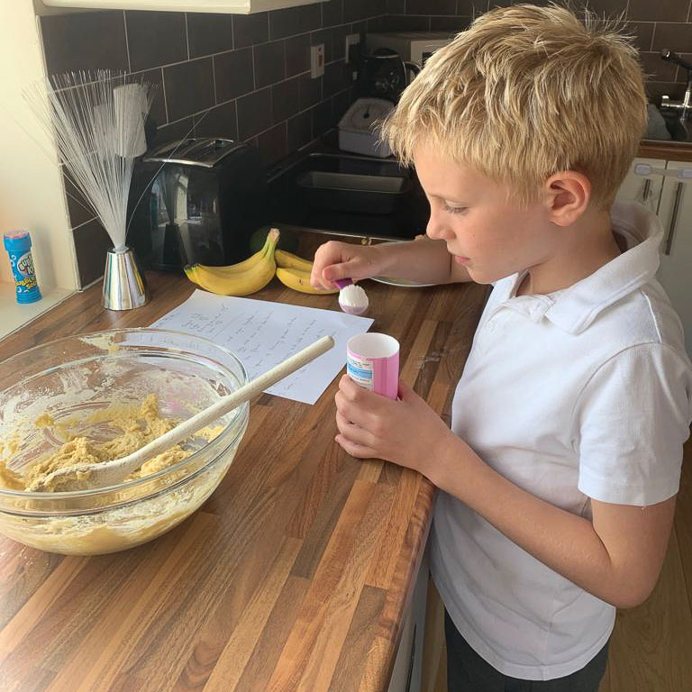 A child is measuring out ingredients for a cake mix. They're adding the baking powder to a large bowl of mixture.