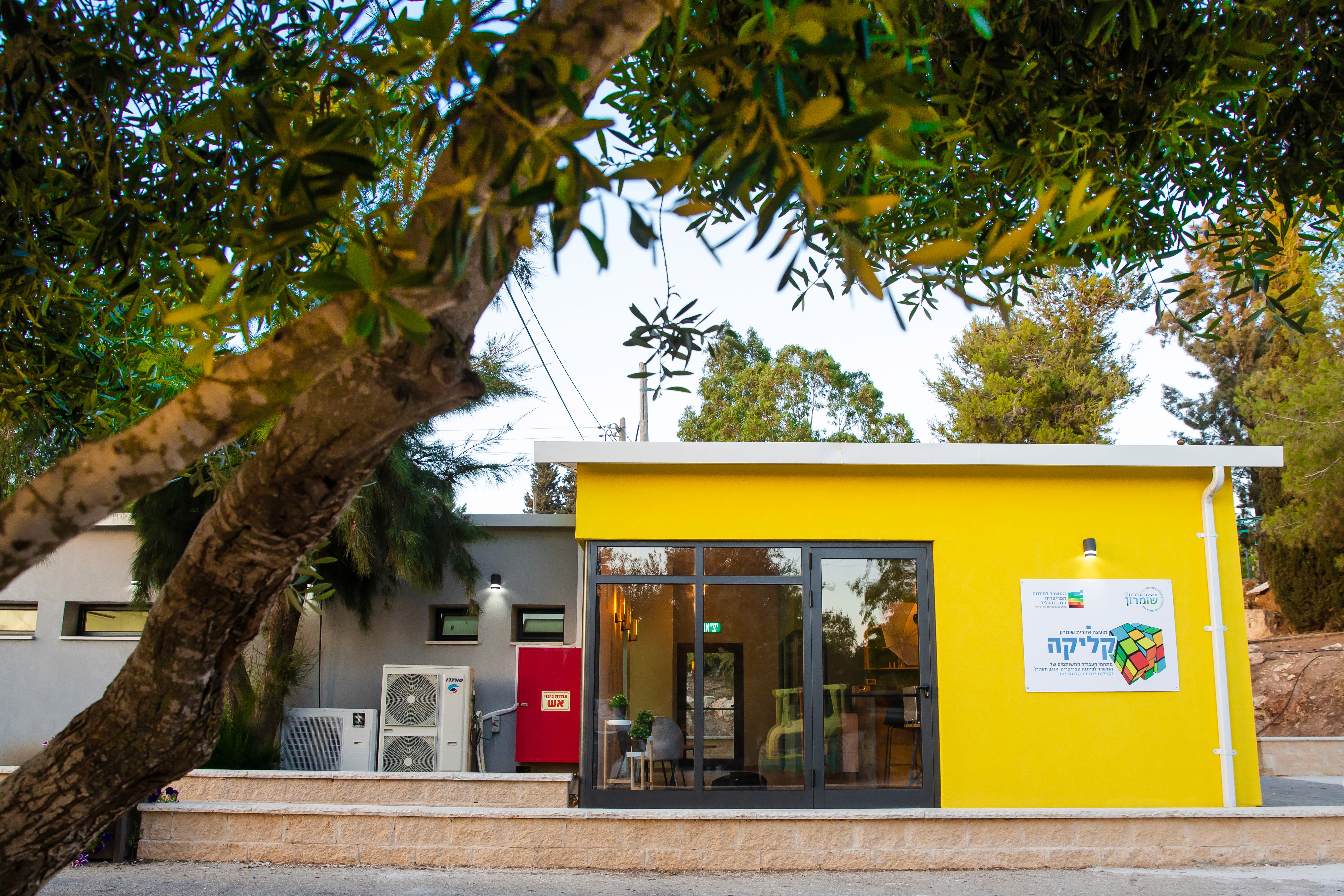 A vibrant yellow building with a modern glass entrance is surrounded by lush greenery and features exterior air conditioning units, set against a backdrop of trees and a clear sky.