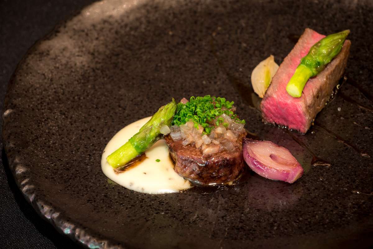 A gourmet plated dish featuring seared meat topped with herbs and a slice of pink tuna with asparagus on a dark plate.