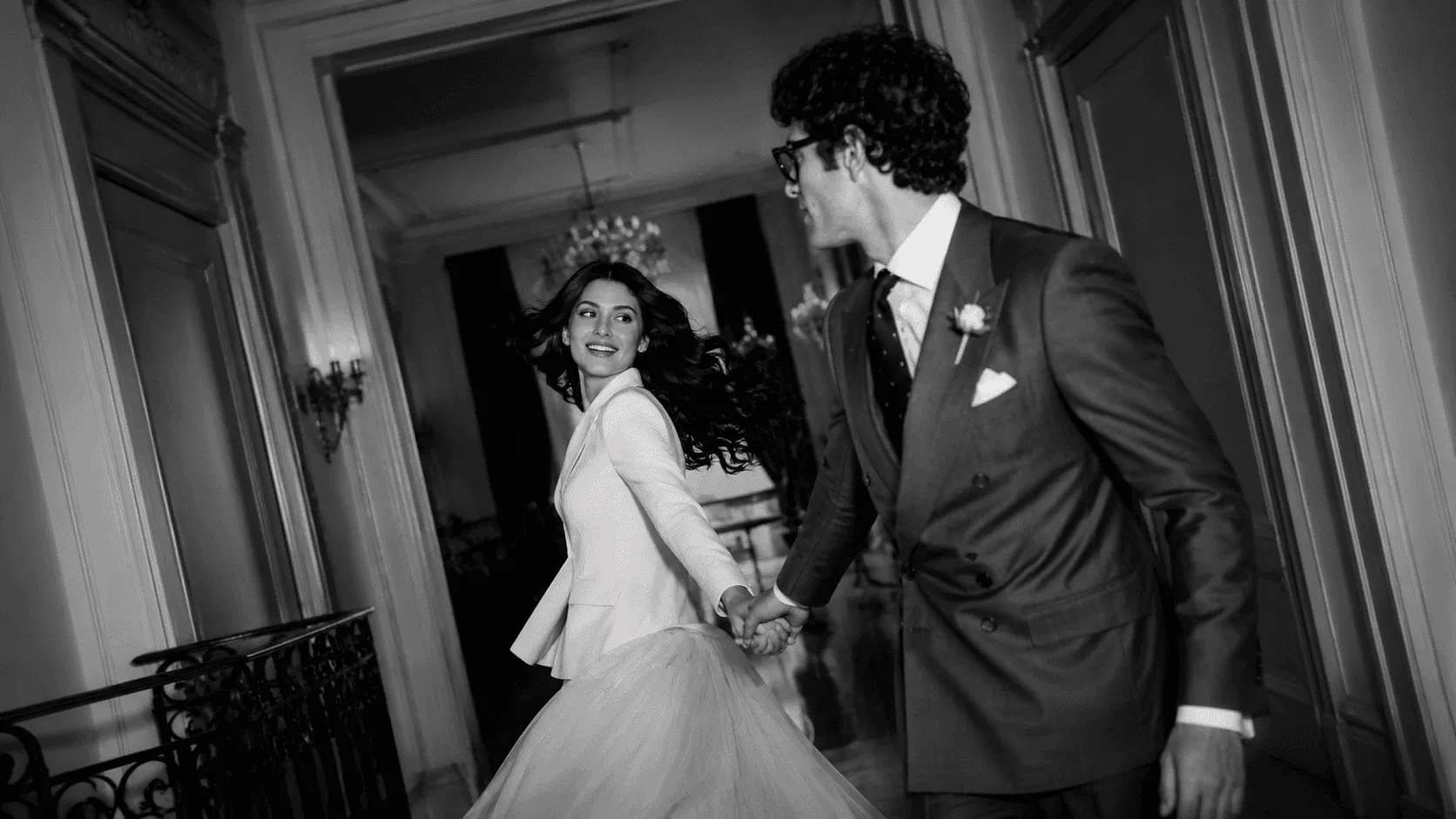 A black and white photo of a joyful couple, with the woman in a flowing wedding dress and the man in a suit, holding hands and running down an elegantly decorated hallway.