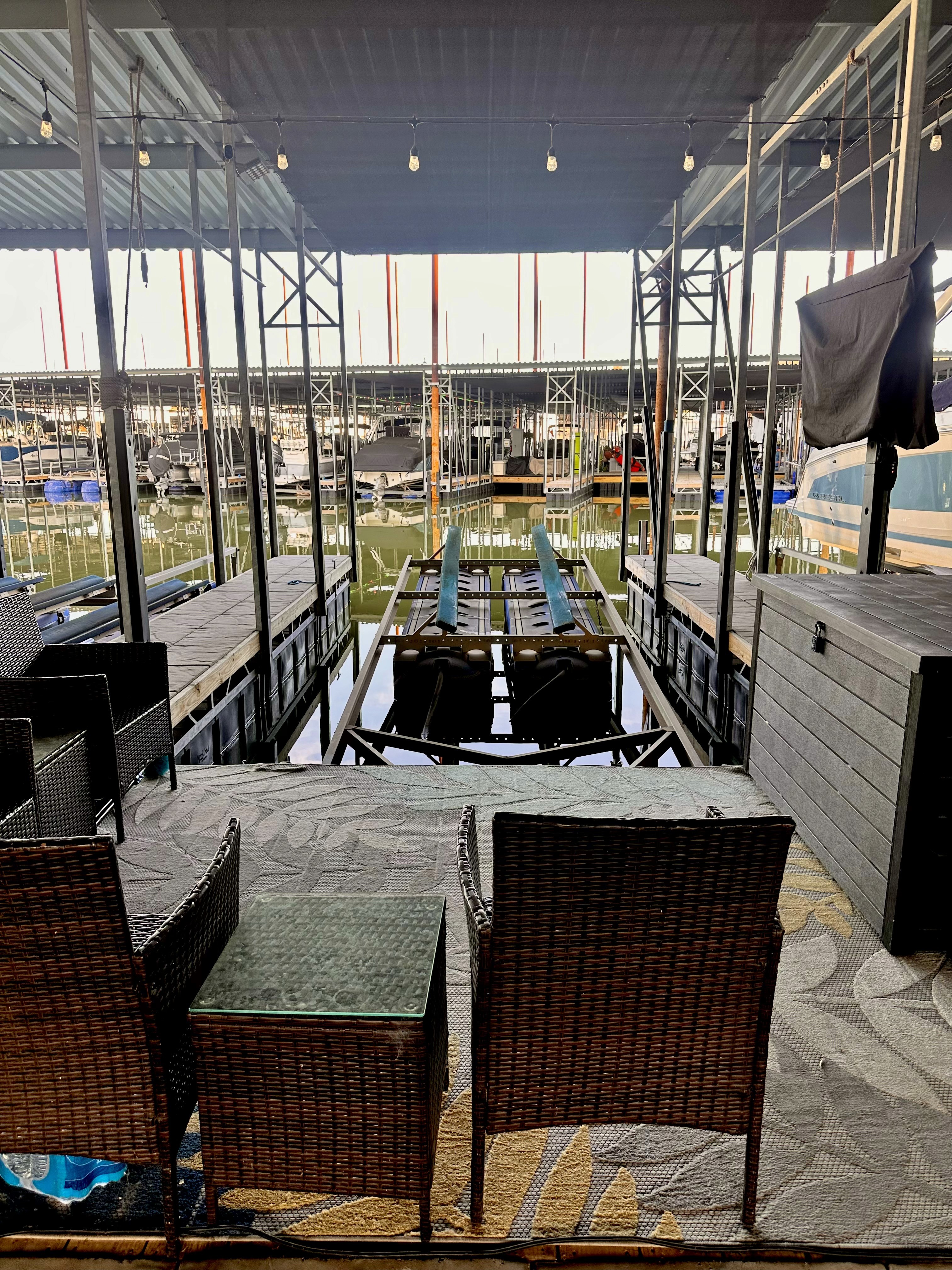 A cozy marina lounge with wicker chairs and a glass-topped table overlooks a row of boats docked in the calm water, framed by steel columns and under a slatted roof, creating a serene waterside retreat.