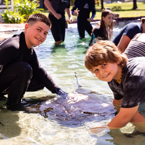 两个微笑的男孩在一个浅水池中抚摸魟鱼，背景中还有其他人参与这个活动。