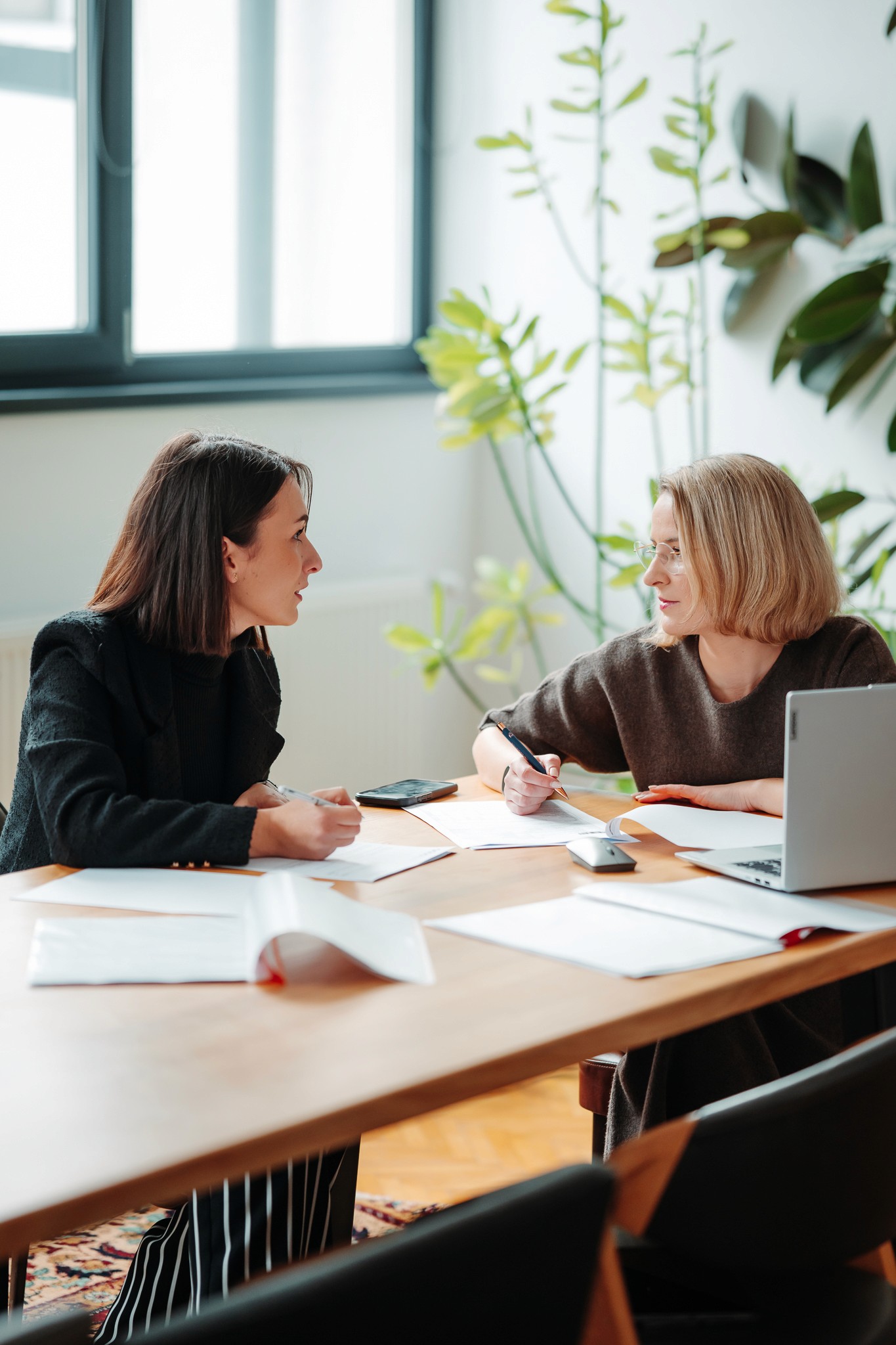 A smiling man with a beard wearing a dark green shirt, sitting at a table with a notebook, coffee cup, and laptop. He gestures with one hand while speaking, with a colorful abstract artwork framed behind him.