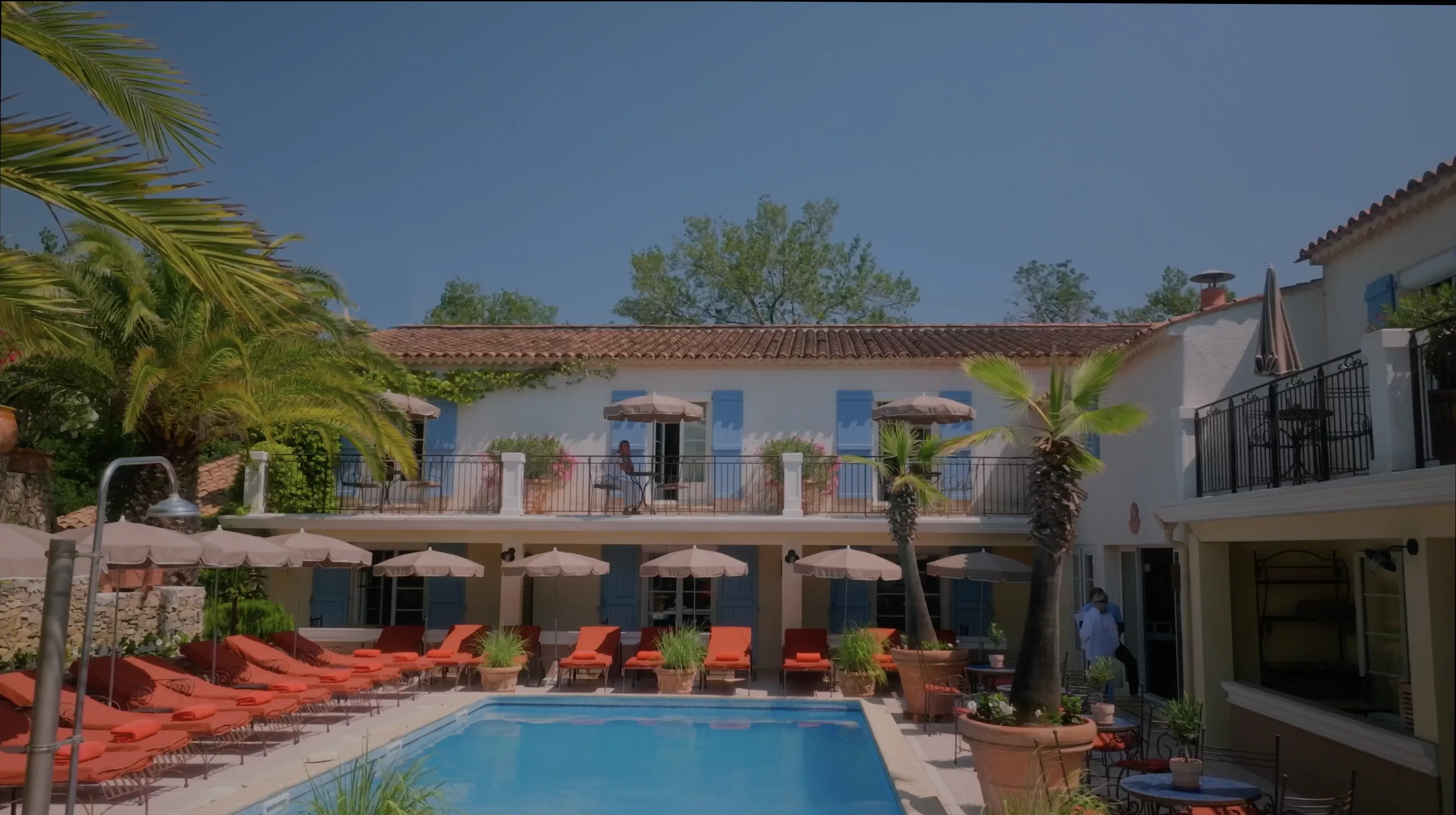 A luxurious Mediterranean-style villa featuring a sparkling blue swimming pool, surrounded by red lounge chairs, tall palm trees, and a two-story building with elegant arched windows and shaded balconies under a clear blue sky.
