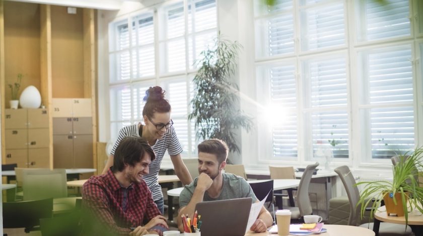 Three people collaborate around a laptop in a bright, modern office filled with natural light and contemporary furnishings, conveying a productive work environment.