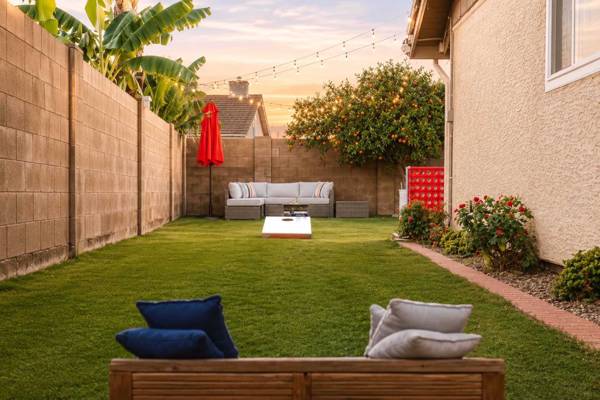 Spacious backyard with lush grass, outdoor sectional seating, and string lights at sunset. Banana trees, roses, and citrus trees line the yard. Yard games like Connect Four included.
