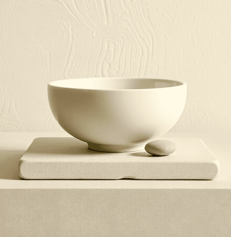 Minimalist white ceramic bowl with a small smooth stone on a beige surface.