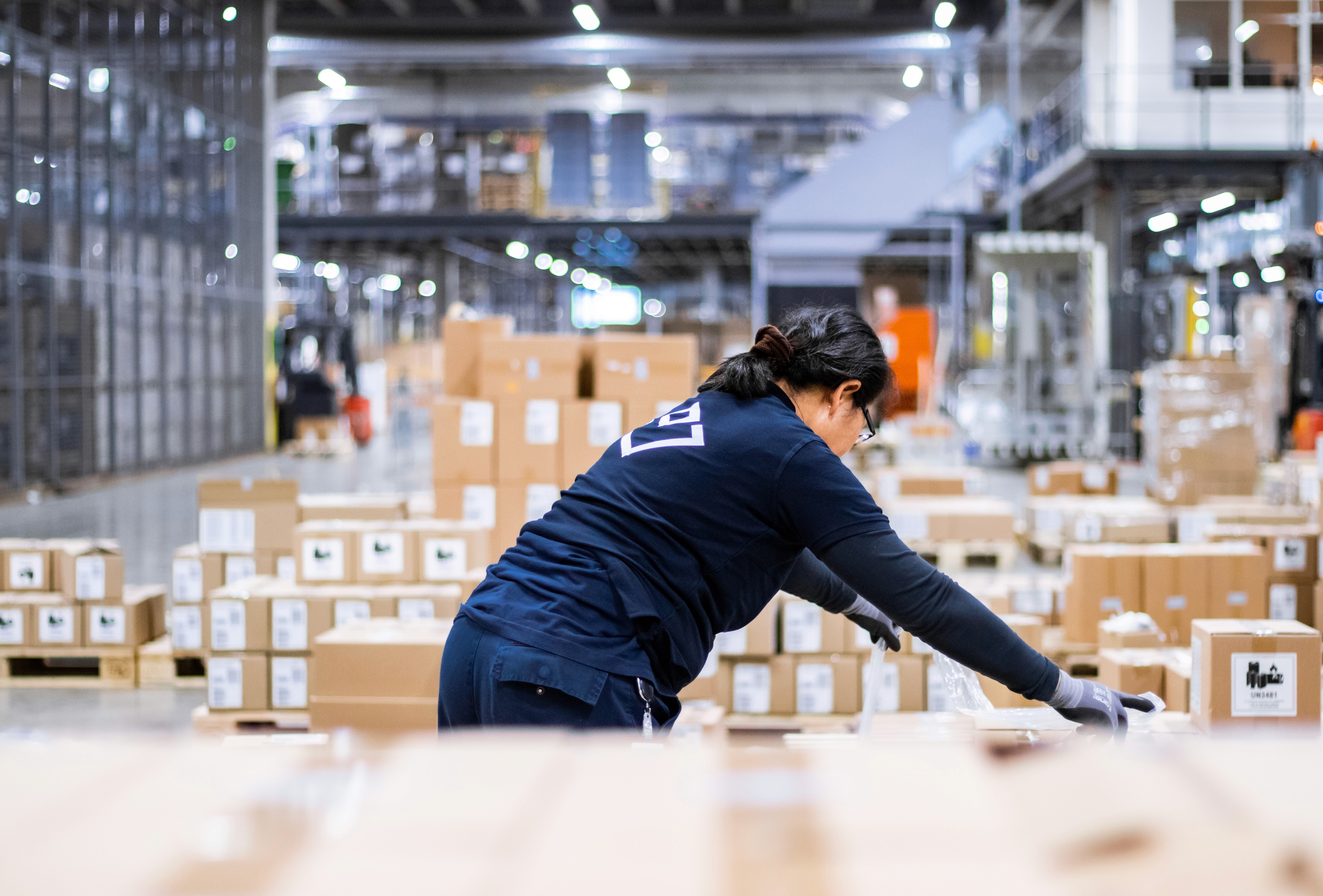 Kvinna sorterar paket i ett lager, klädd i blått och svarta handskar, omgiven av kartonger.