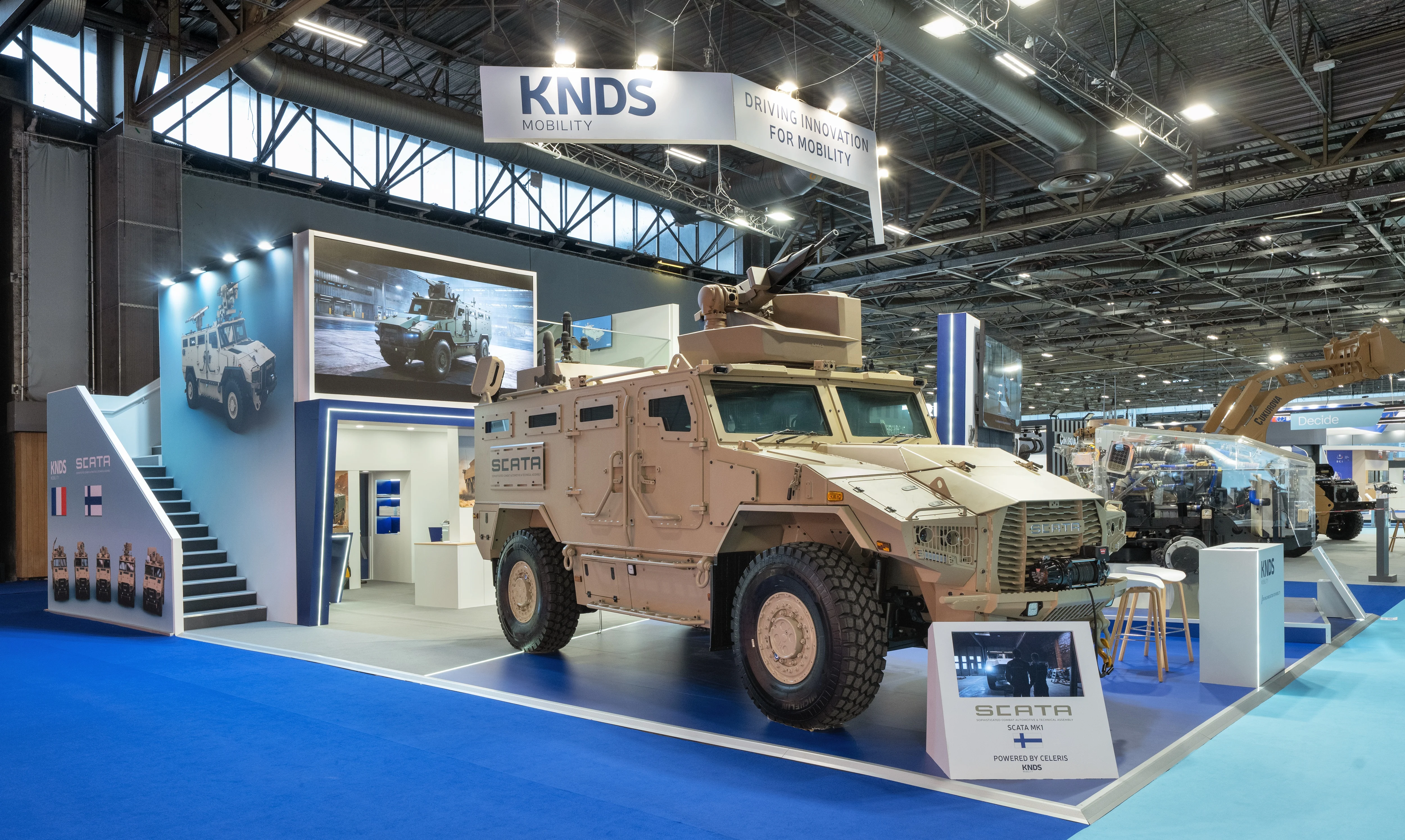 Stand KNDS Mobility conçu et réalisé par Expace à Eurosatory 2026 – espace sur-mesure défense avec blindé SCATA, châssis Celeris écorché, bandeau suspendu « Driving Innovation for Mobility » et lounge à l'étage.