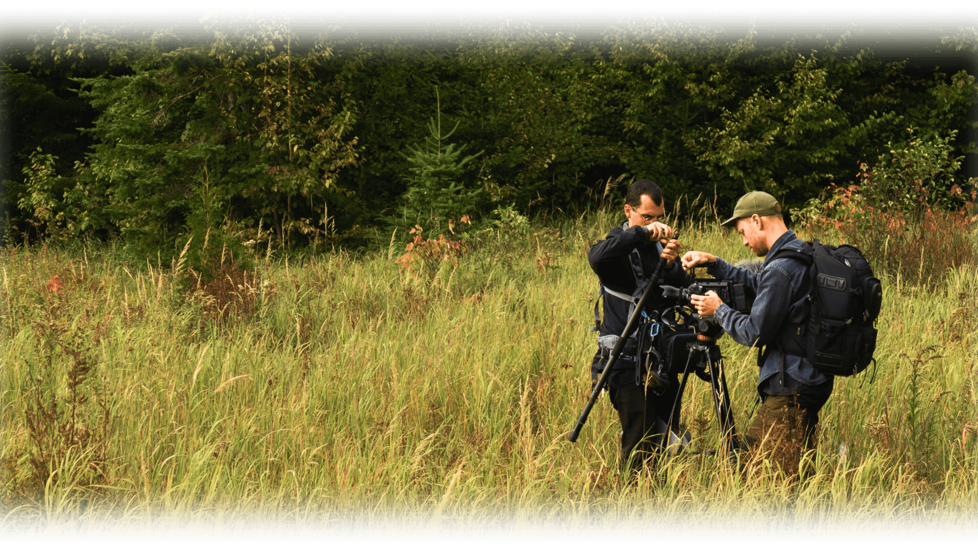 Deux personnes ajustant du matériel audio et vidéo sur un trépied au milieu d’une prairie, entourées d’herbes hautes et d’une forêt dense.